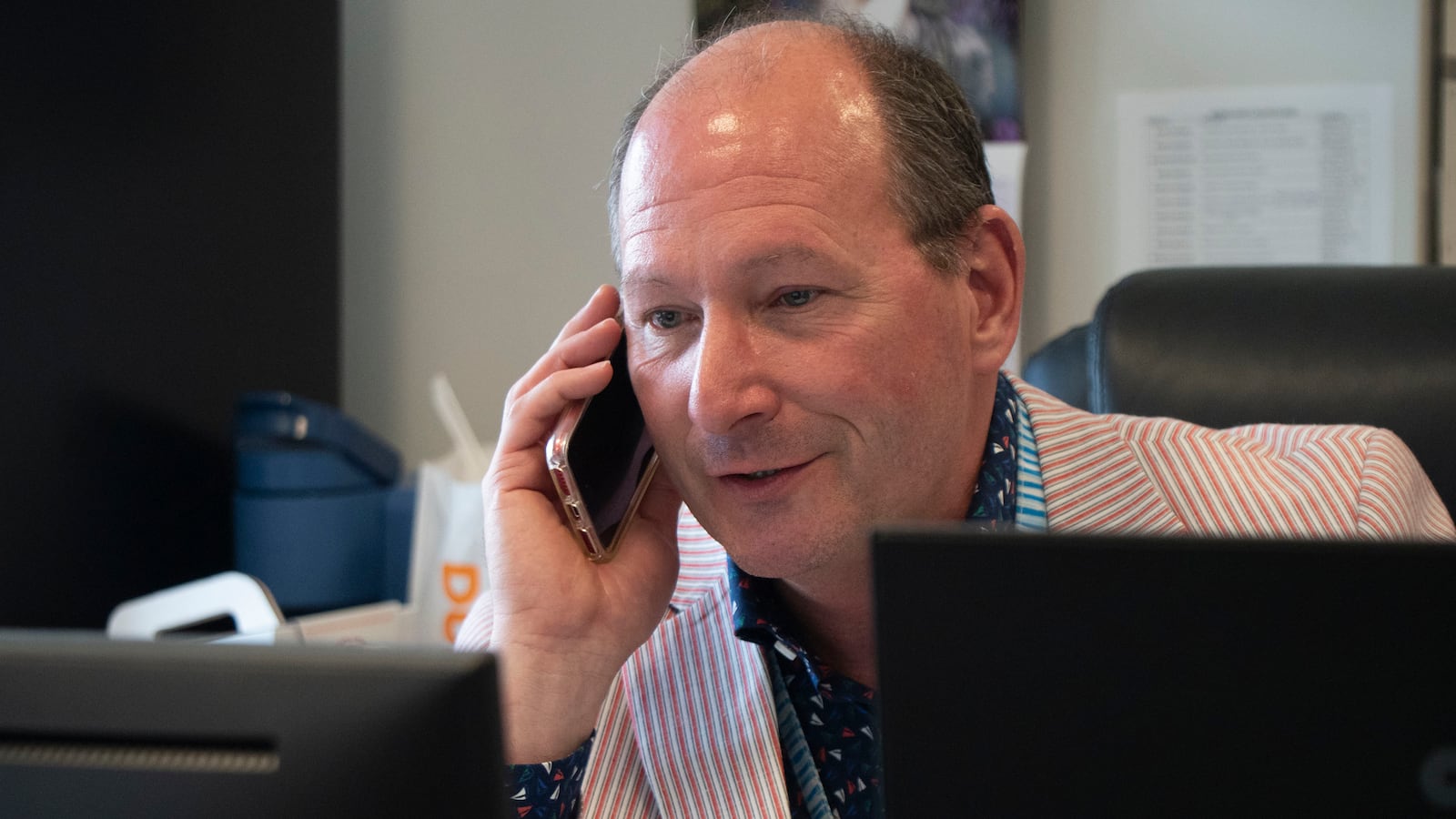 A man wearing a red and white striped suit sits at a desk behind computer screens and holds a phone to his ear.