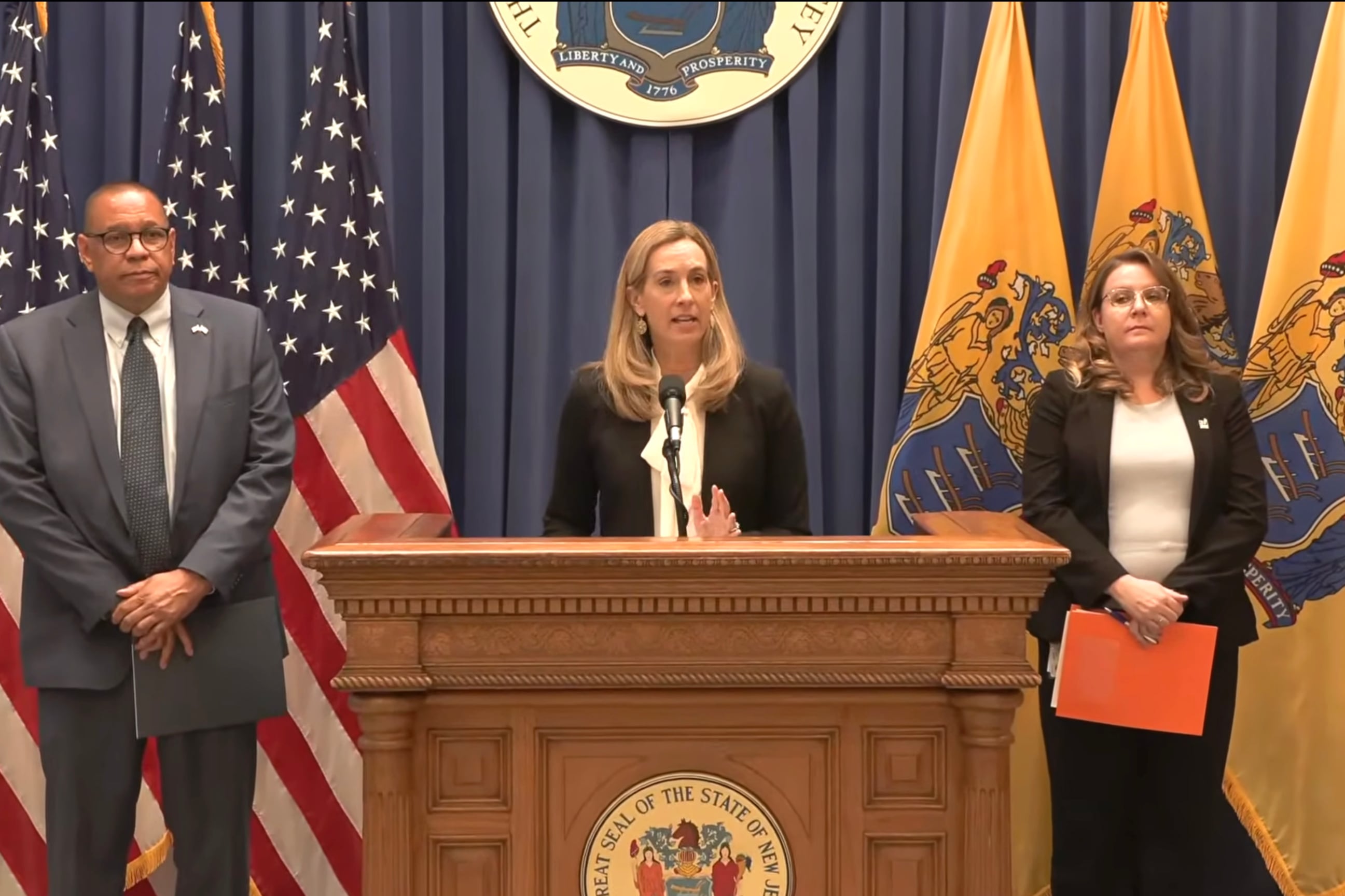 A screen grab of a youtube video of one white woman in the middle speaking into a small microphone on a wooden podium with two people standing on either side of her in front of six flags.
