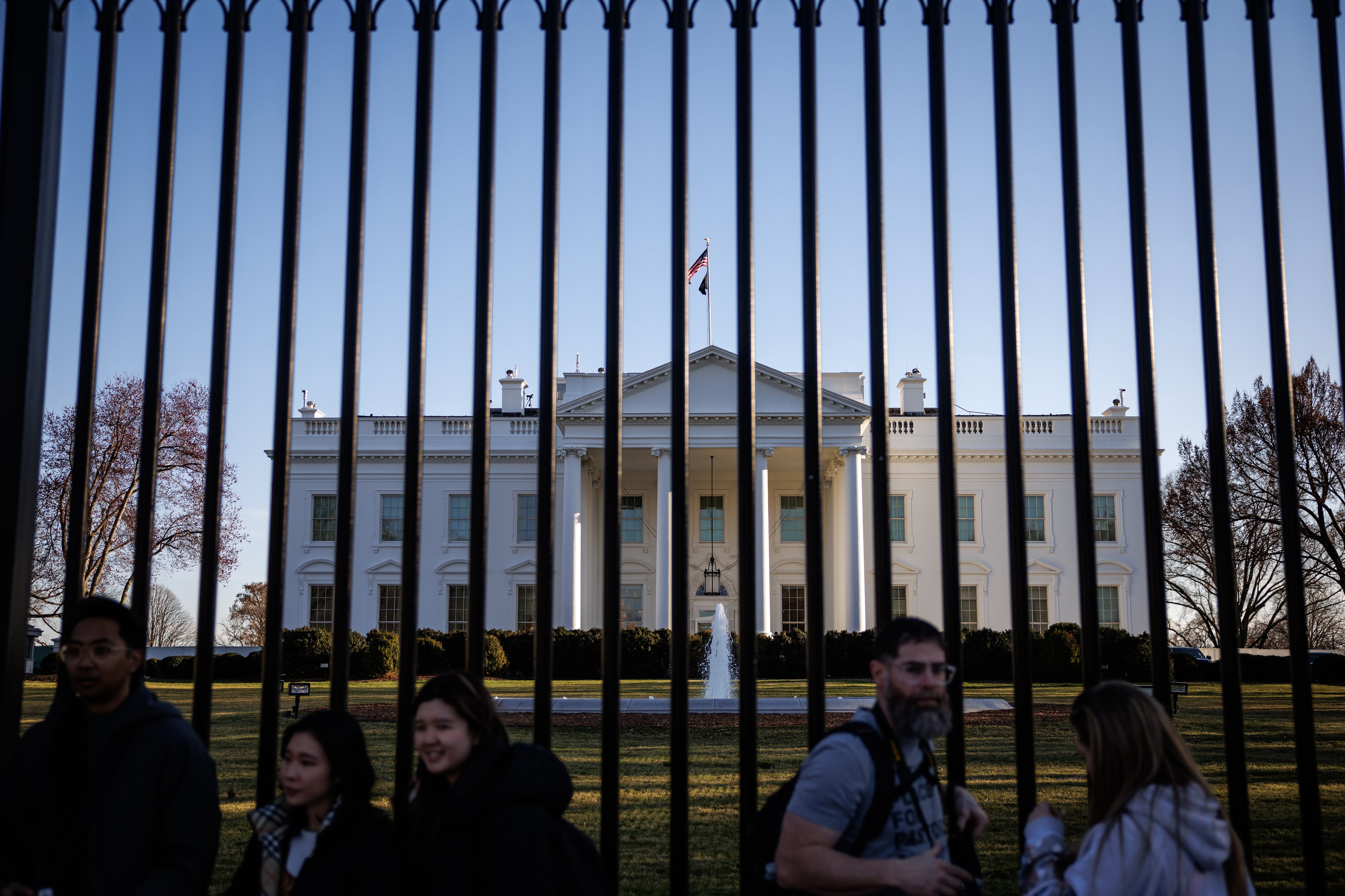 A tall fence with the White House in the background and five people standing in front of the fence.