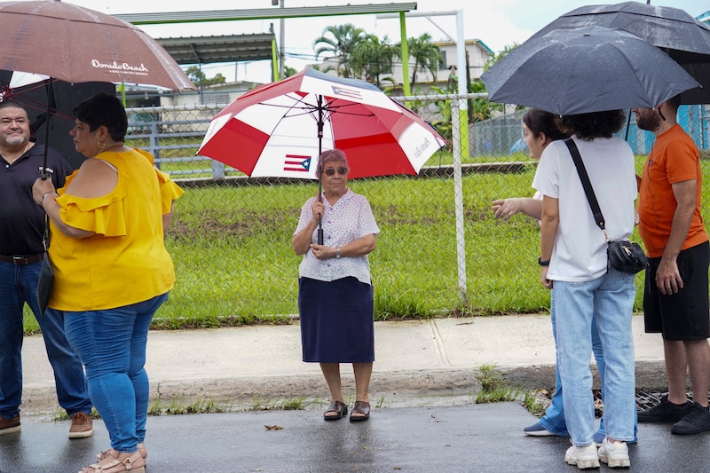 A woman holding a red and white umbrella stands in a line of people outside.