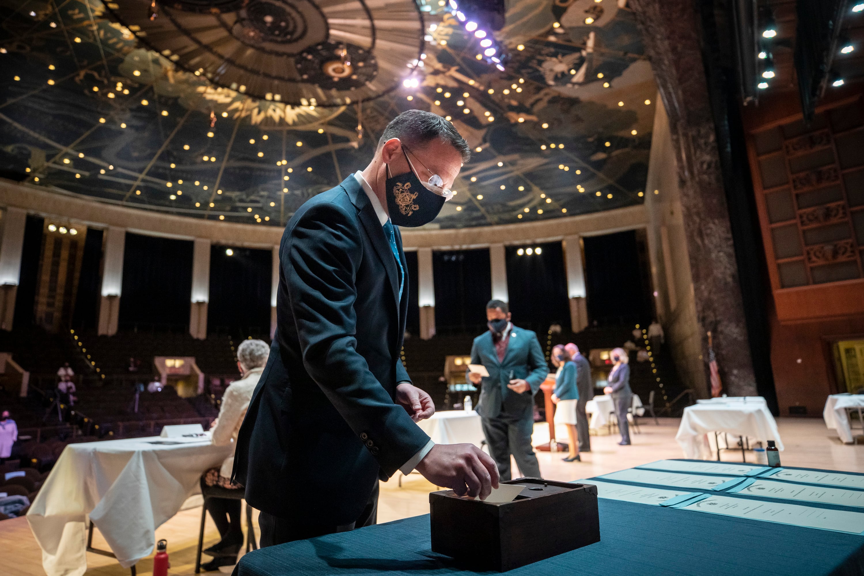 A man wearing a dark suit and a face mask places a ballot into a small black box to cast a vote in a large room with a dome ceiling and people standing in the background.