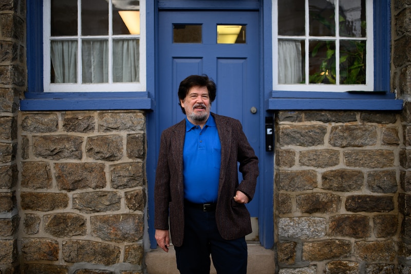 A man with dark brown hair and wearing a bright blue shirt under a jacket poses for a photograph in front of a blue door.