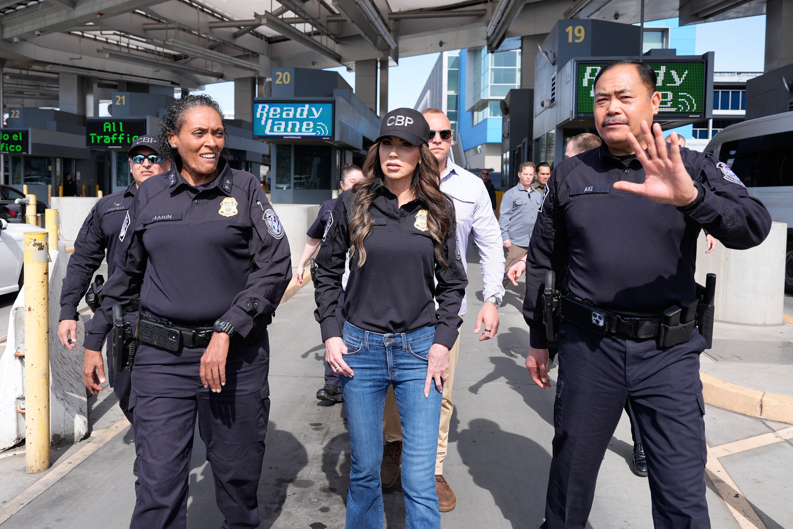 Three people in uniforms walk in a line facing the camera while a group of people walk behind them.