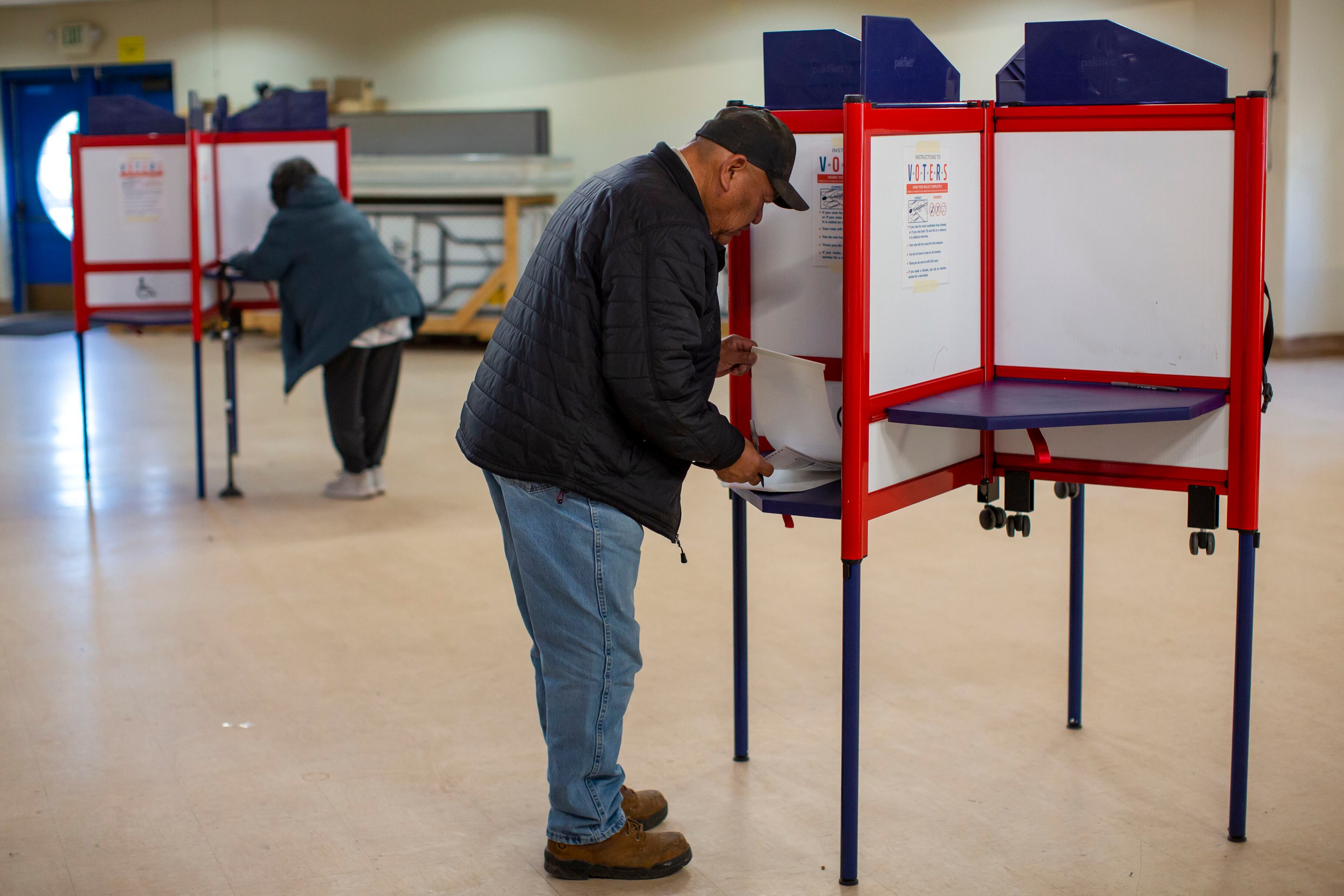 Two people stand in front of ballot secrecy stands while voting in a room.