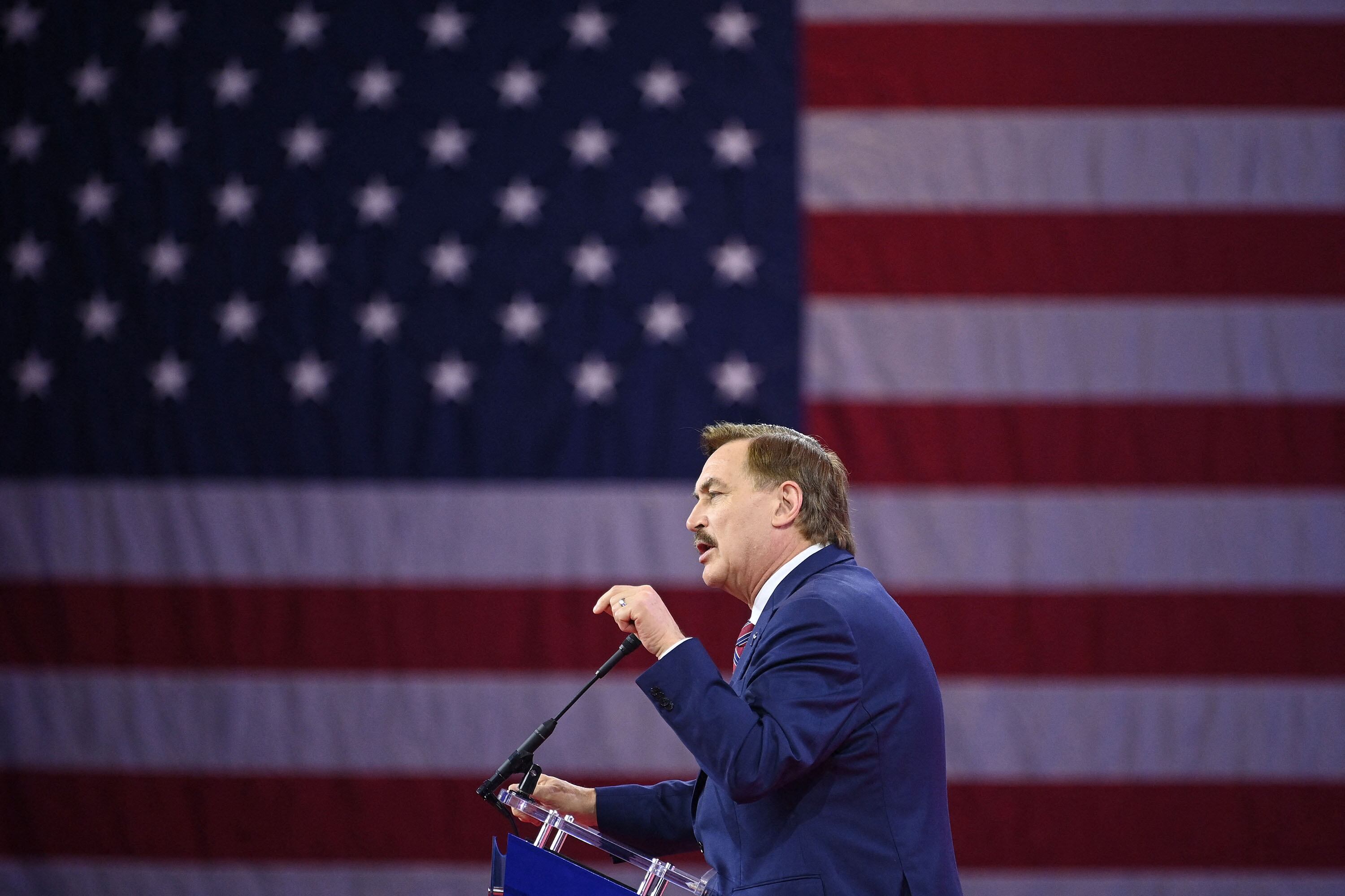 A profile photo of a man with short brown hair and wearing a dark blue suit speaks at a podium with a giant American flag taking up the whole background.