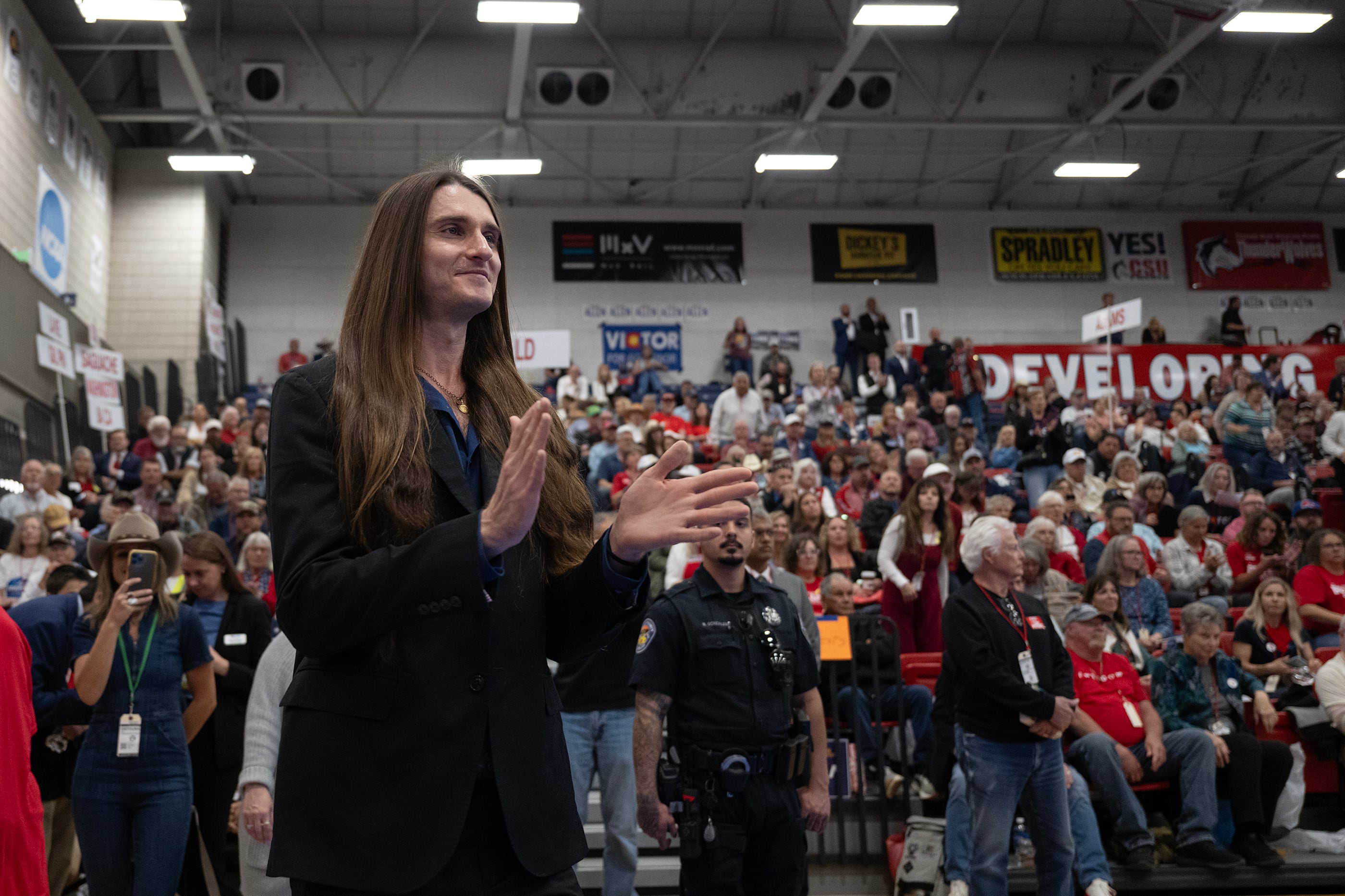 A white man with long blonde hair stands clapping with a large group of people standing behind him clapping.