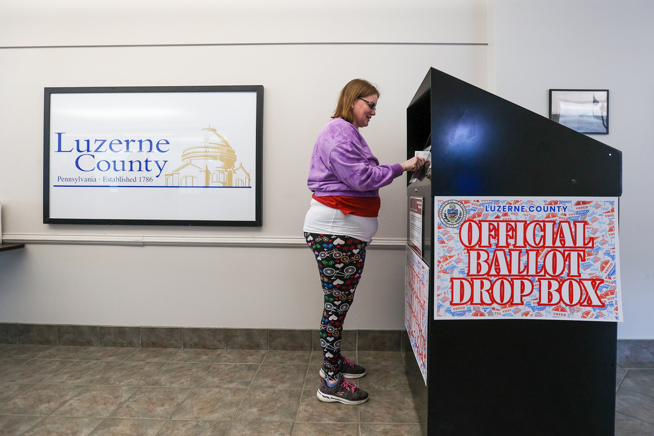 A photograph of a white woman placing a ballot into a large metal box with a sign on the left on the back wall that reads "Luzerne County"