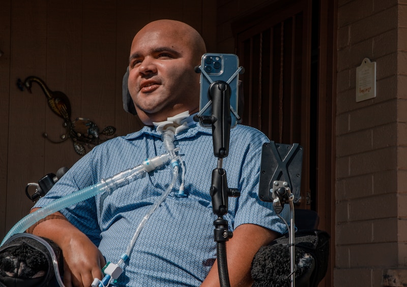 A man wearing a blue shirt with medical tubes going down the front sits in a motorized chair in front of a house.