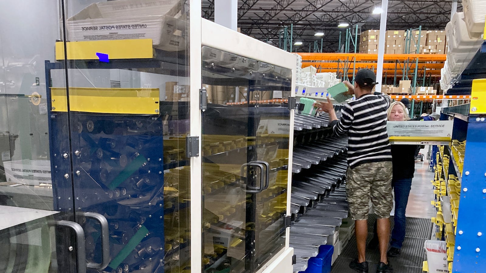 A person wearing a striped shirt and cargo shorts stands next to a giant machine that sorts voting ballots.