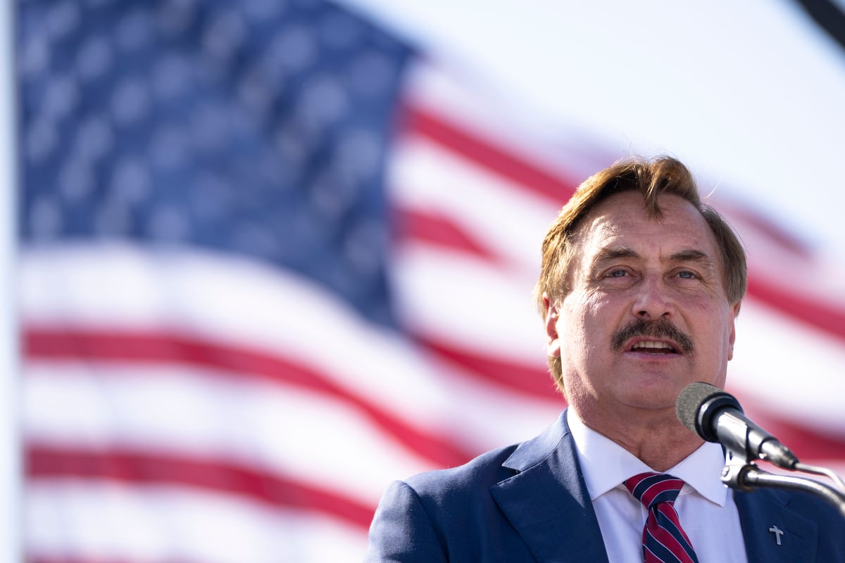 A man with brown hair and a mustache stands at a microphone, with a blurry American flag in the background.