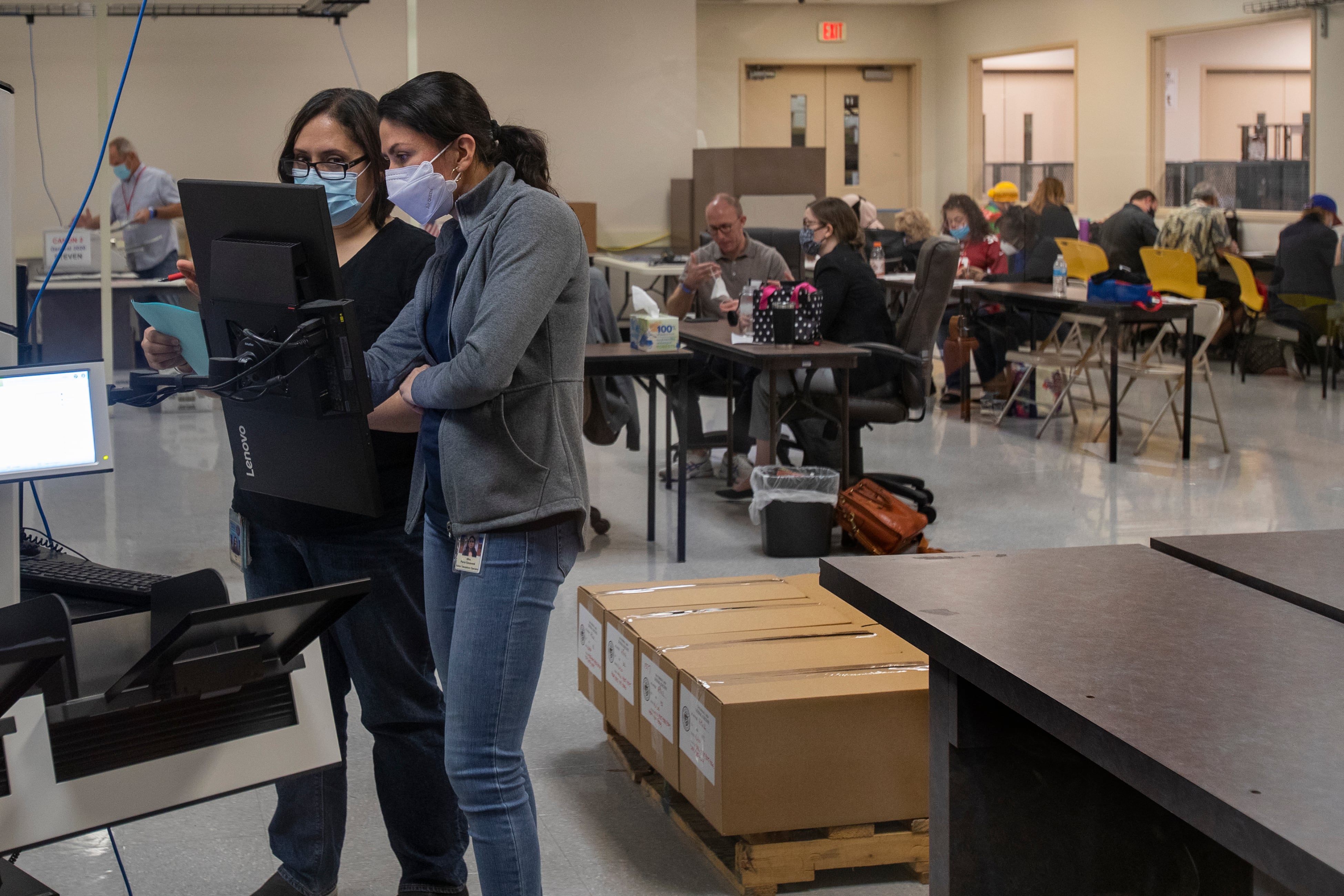 Two women examine a screen in a room with other workers at tables