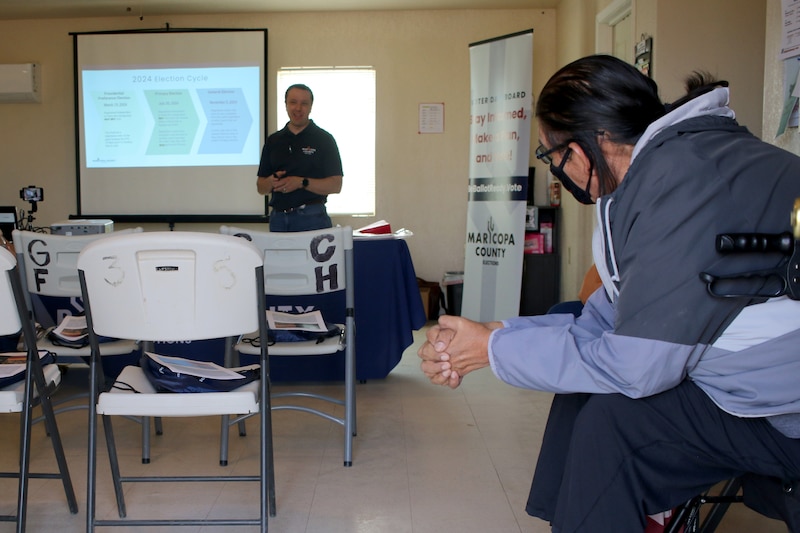 A person stands in the back of the room next to a projector screen while a person sits on a chair looking their way in the foreground.