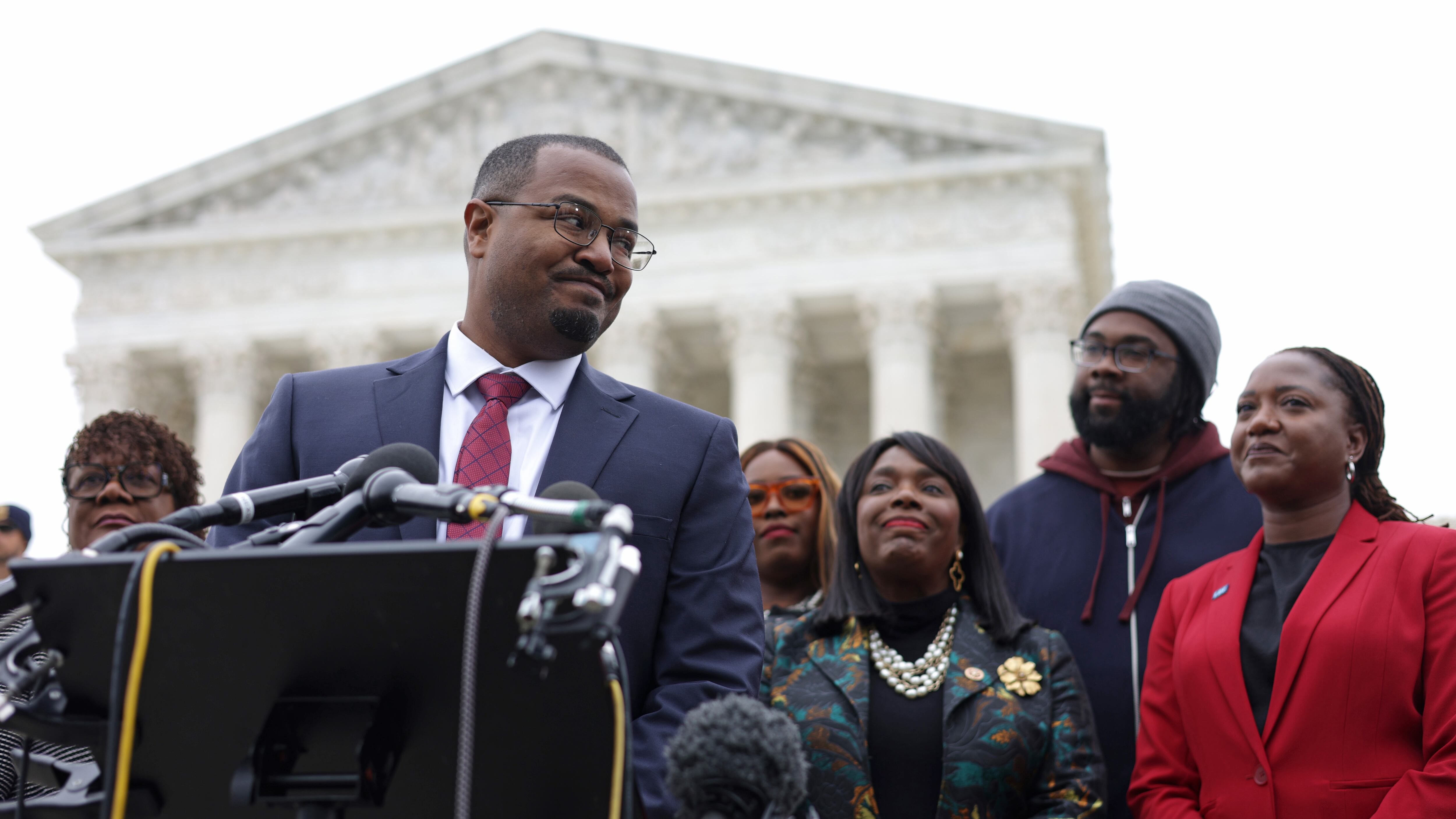 A man wearing glasses stands behind a podium with several microphones on it with several people standing behind him and the Supreme Court in the background.