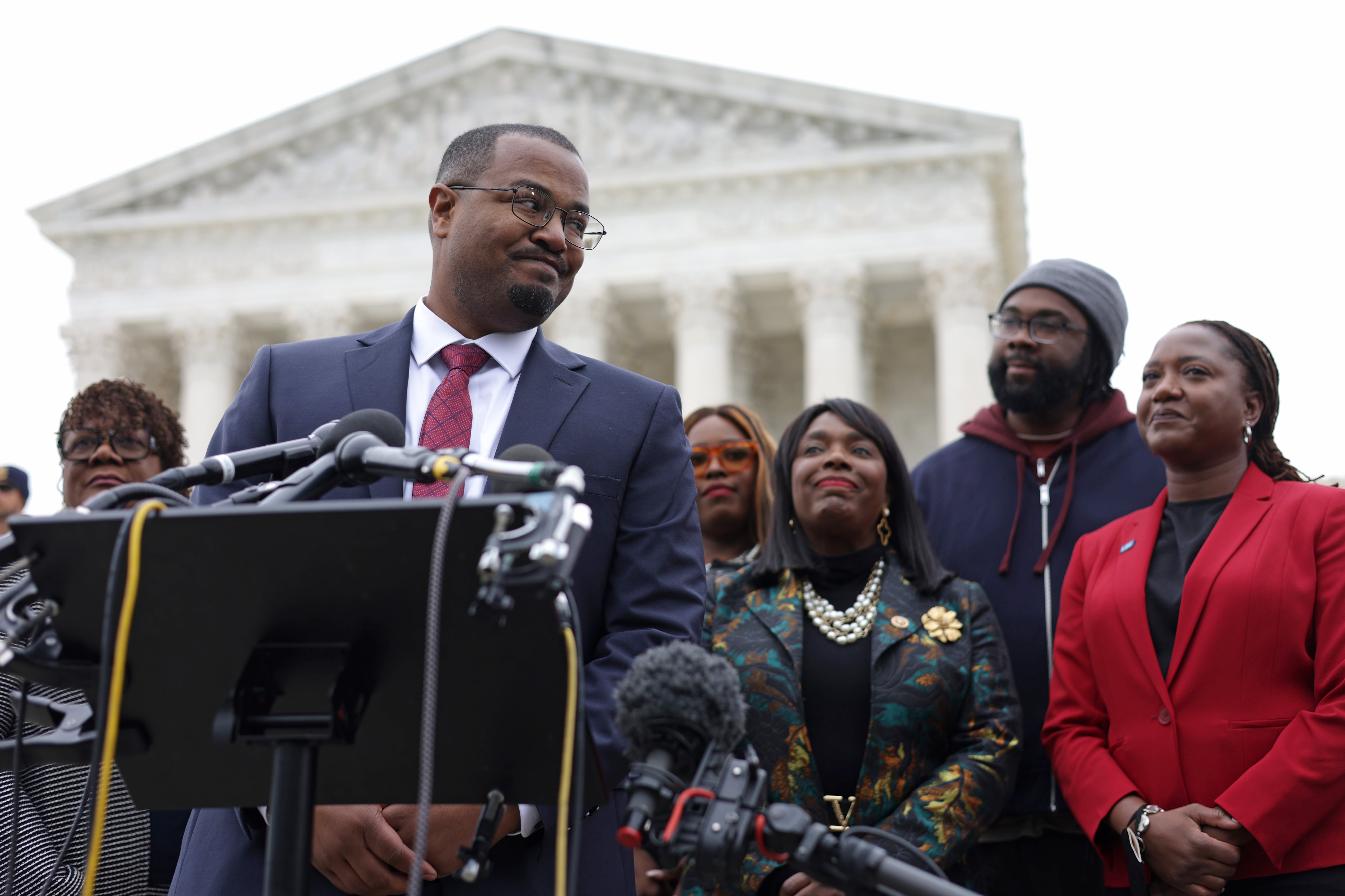 A man wearing glasses stands behind a podium with several microphones on it with several people standing behind him and the Supreme Court in the background.