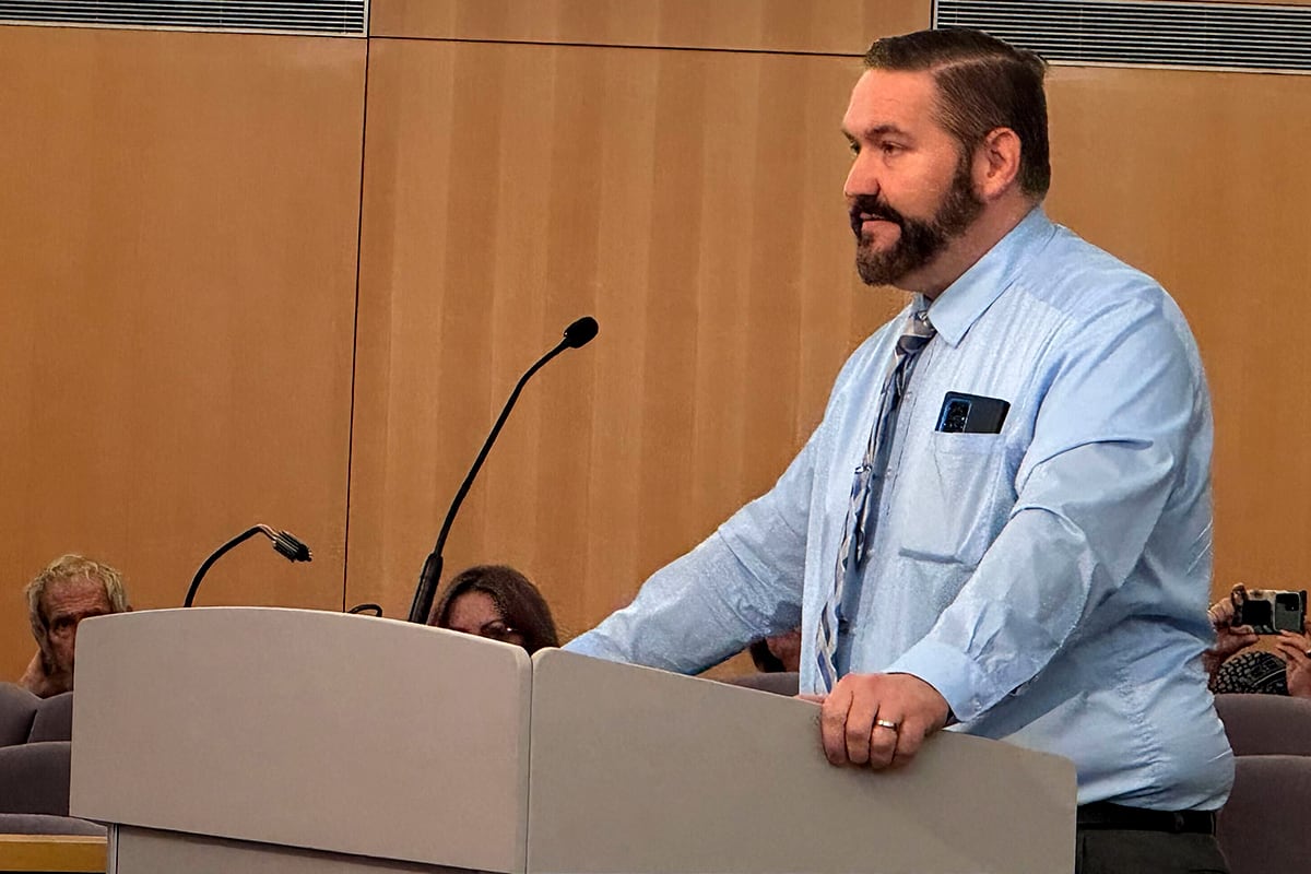 A photograph of a white man in a dress shirt standing behind a wooden podium in a conference room.