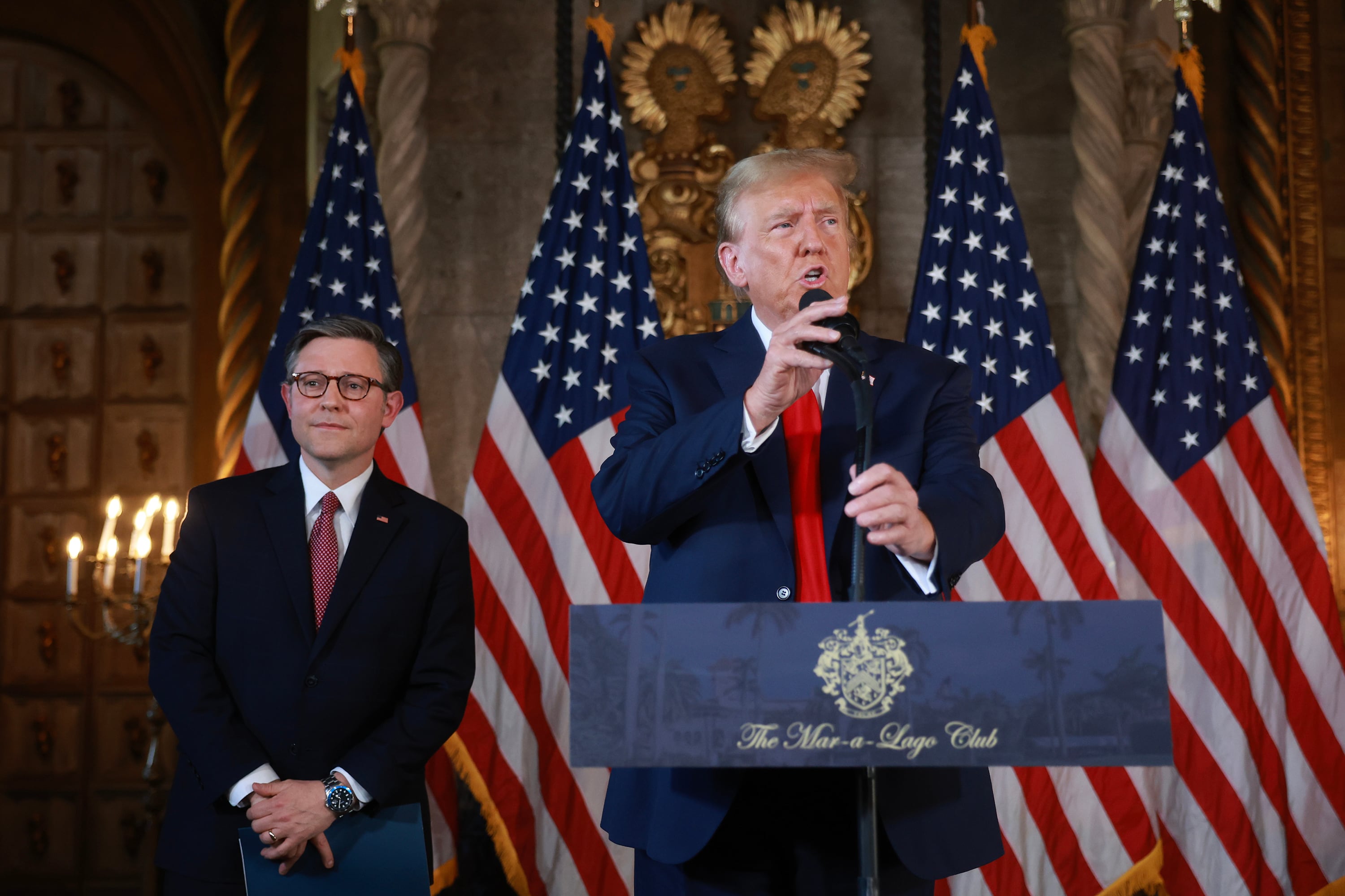 A man wearing a dark suit speaks from a podium with another main in a suit stands next to American flags in the background.