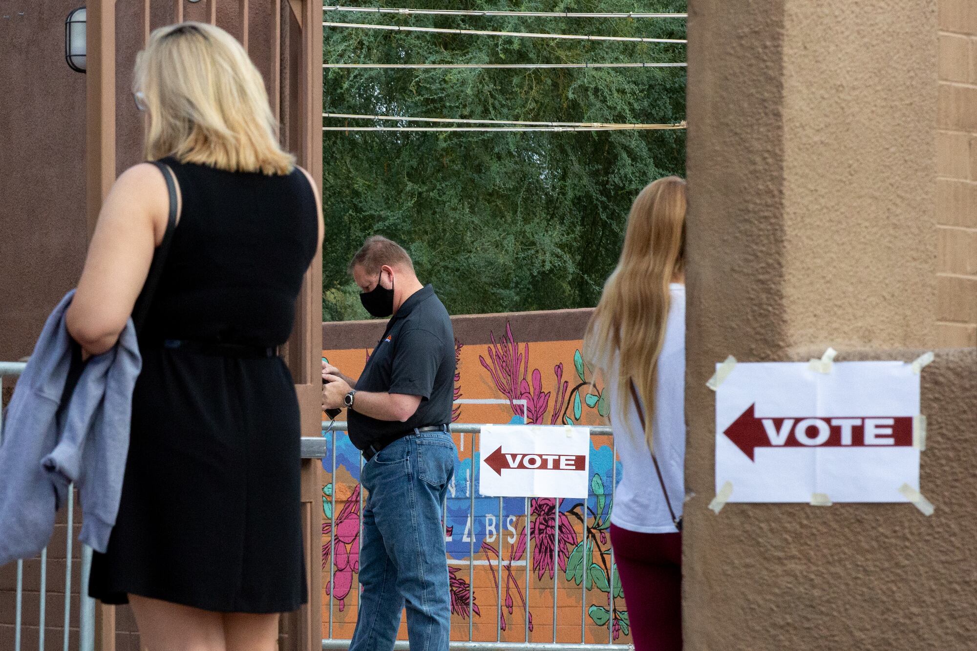 Two women with their backs to the camera and a masked man wait against beige pillars plastered with signs reading “vote.”