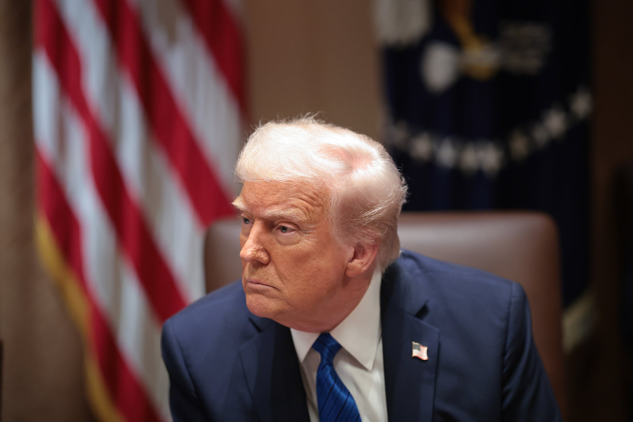 President Donald Trump sits in a chair looking out of the frame with an American flag in the background.
