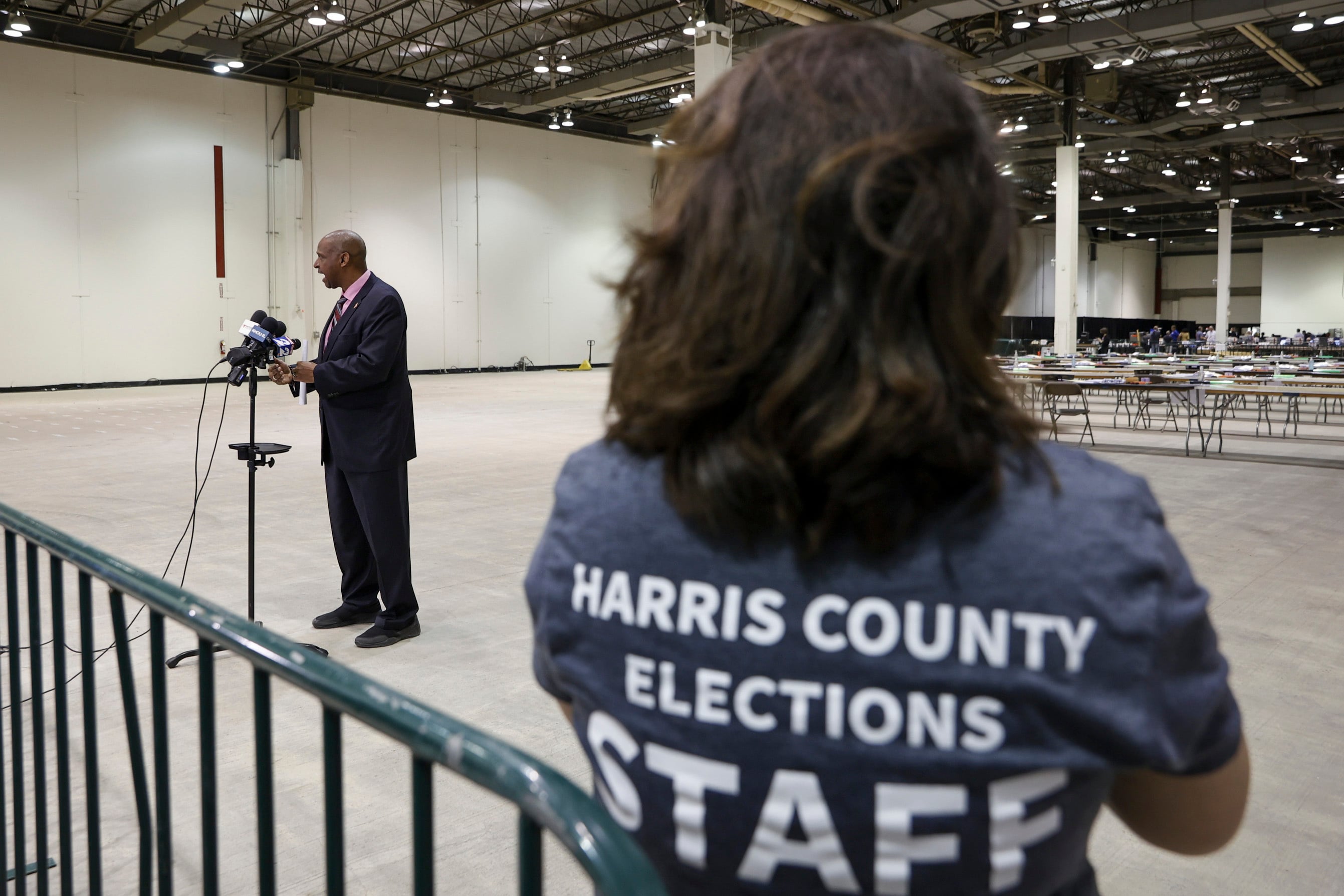 A woman watches a man standing in a suit speak into several microphones