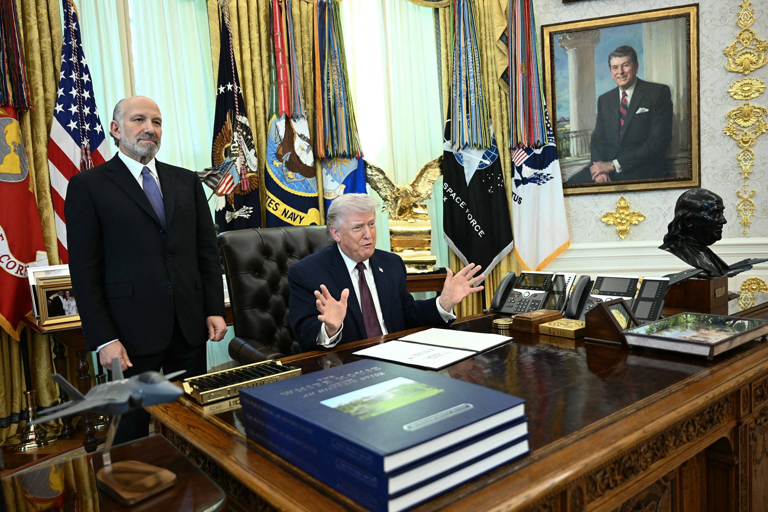 A photograph of two white men in suits in the oval office. President Trump in a blue suit and red tie sits at his desk while the other man stands next to him.