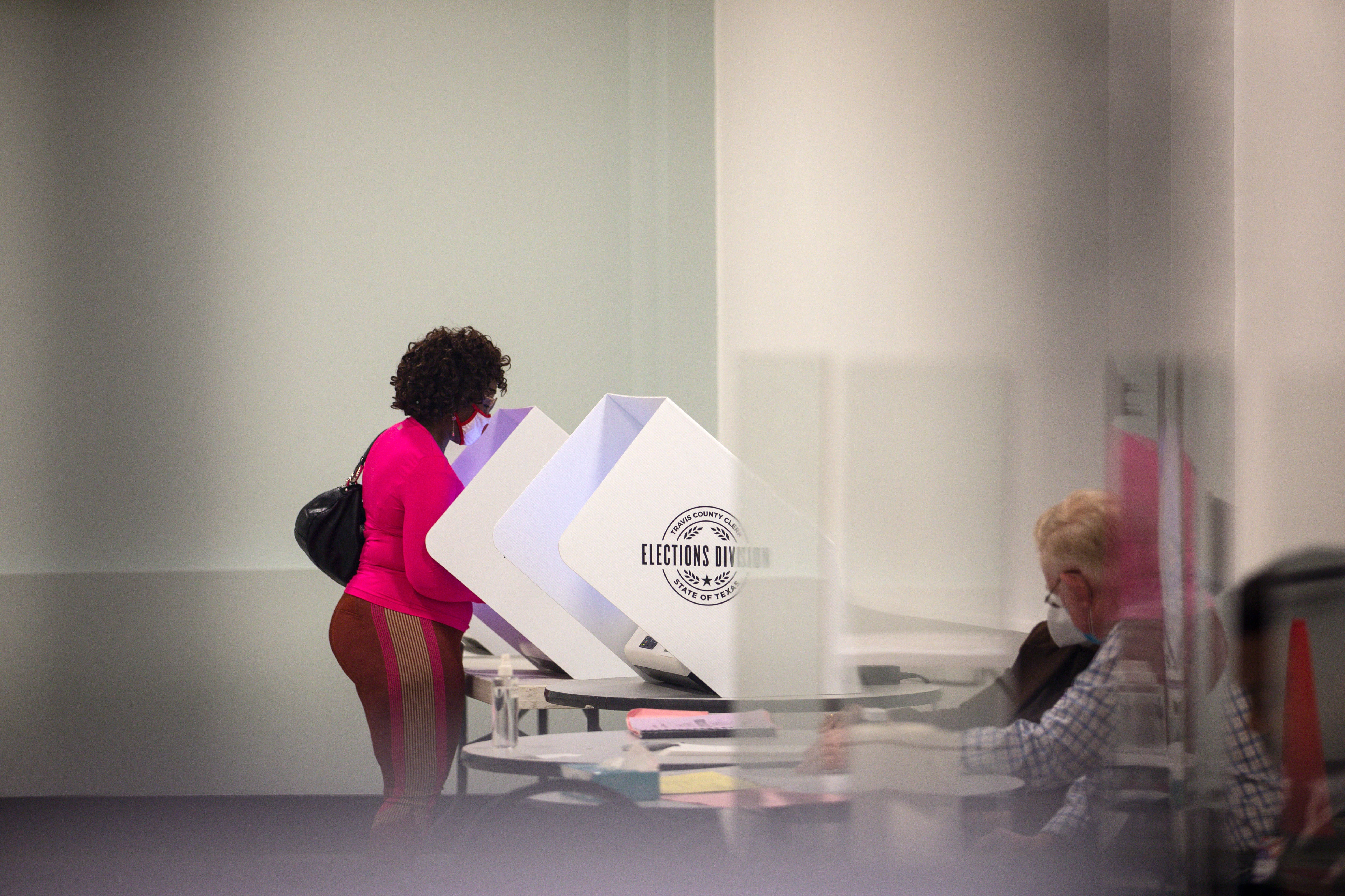 A woman in a pink blouse votes in a primary election as election workers sit behind glass partitions.