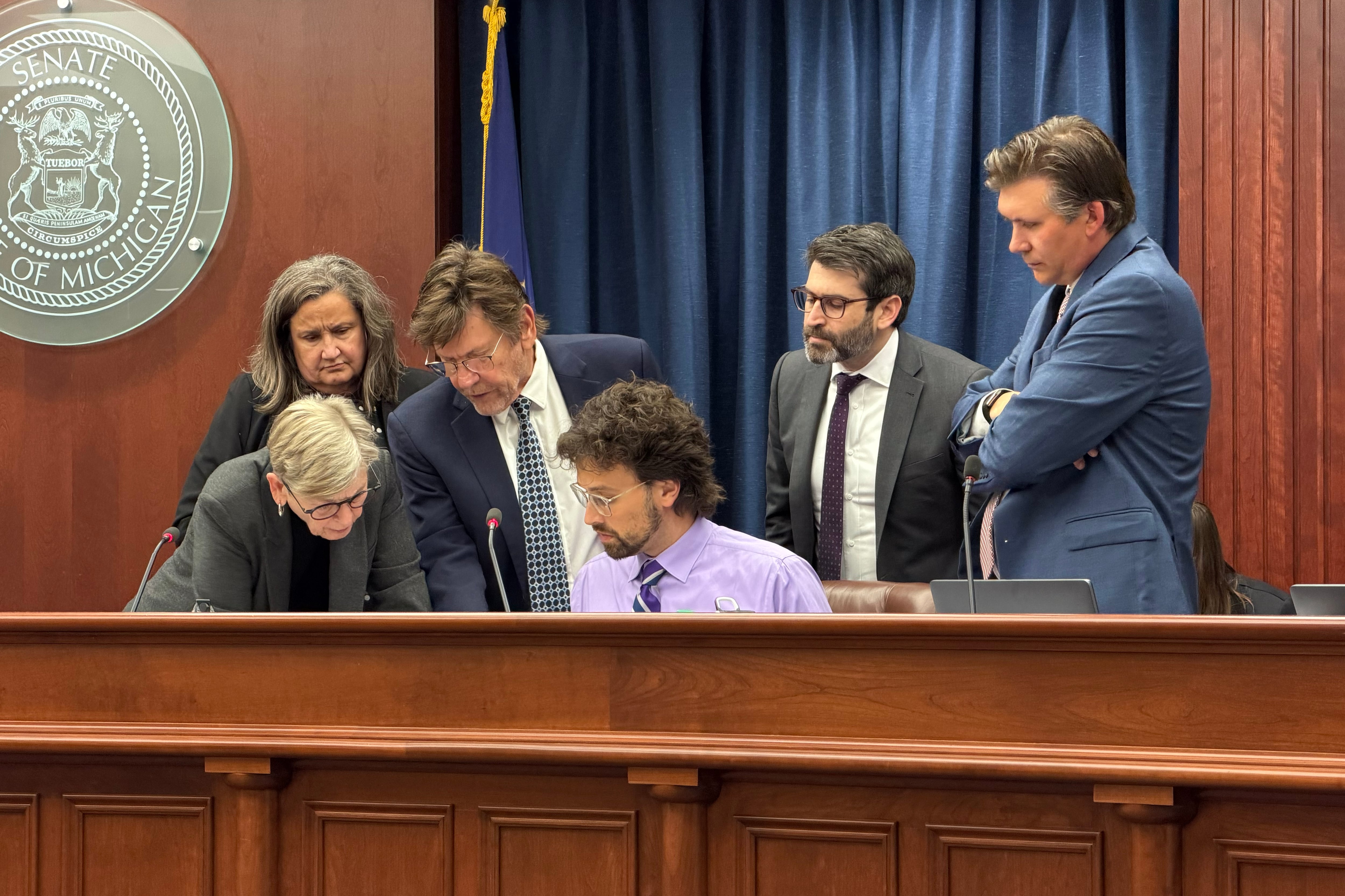 Six white adults in suits stand around a person sitting at a wooden table in a state conference room.