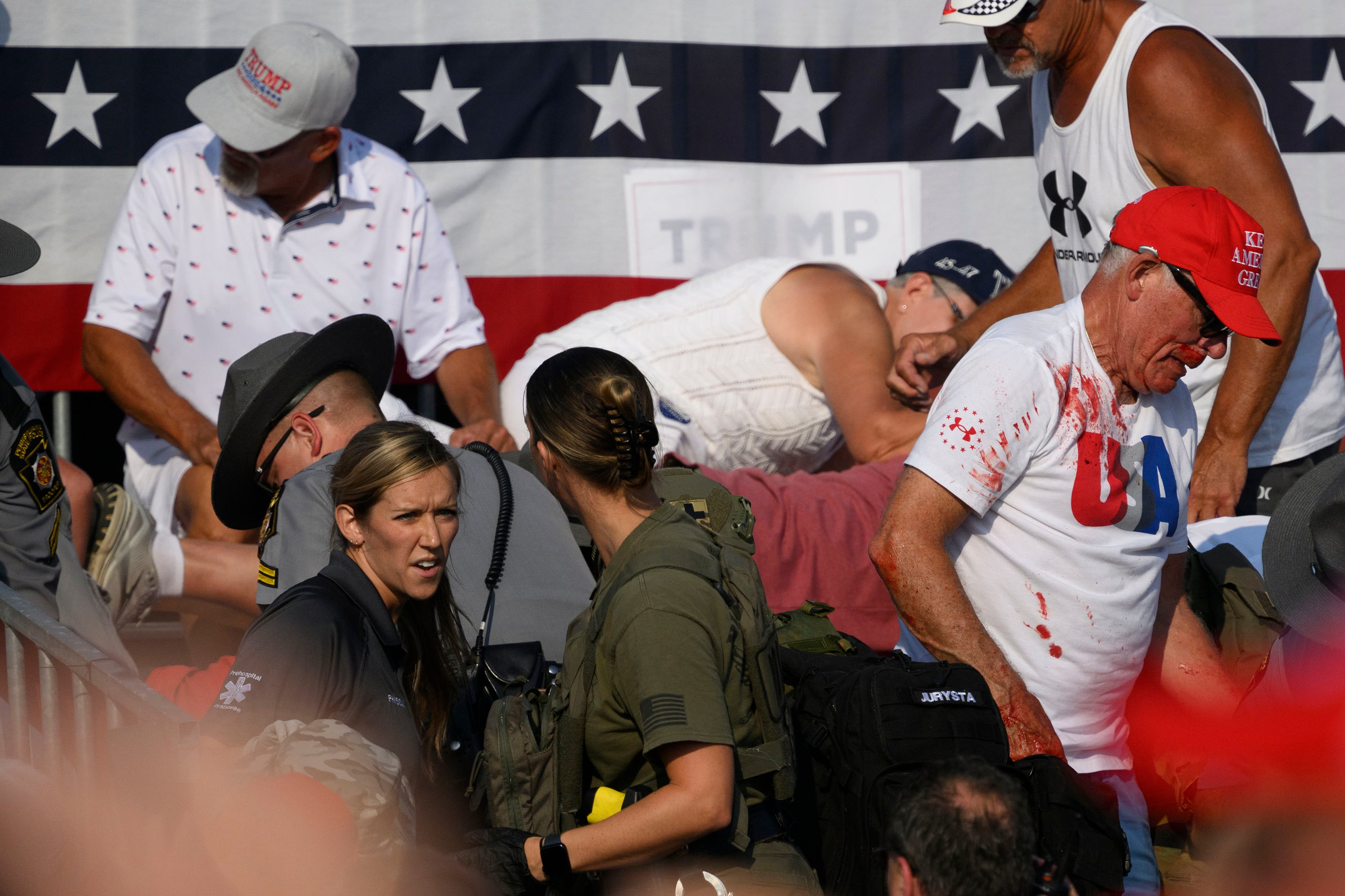 A crowd of people duck and move after hearing gunshots with a large red, white and blue banner in the background.