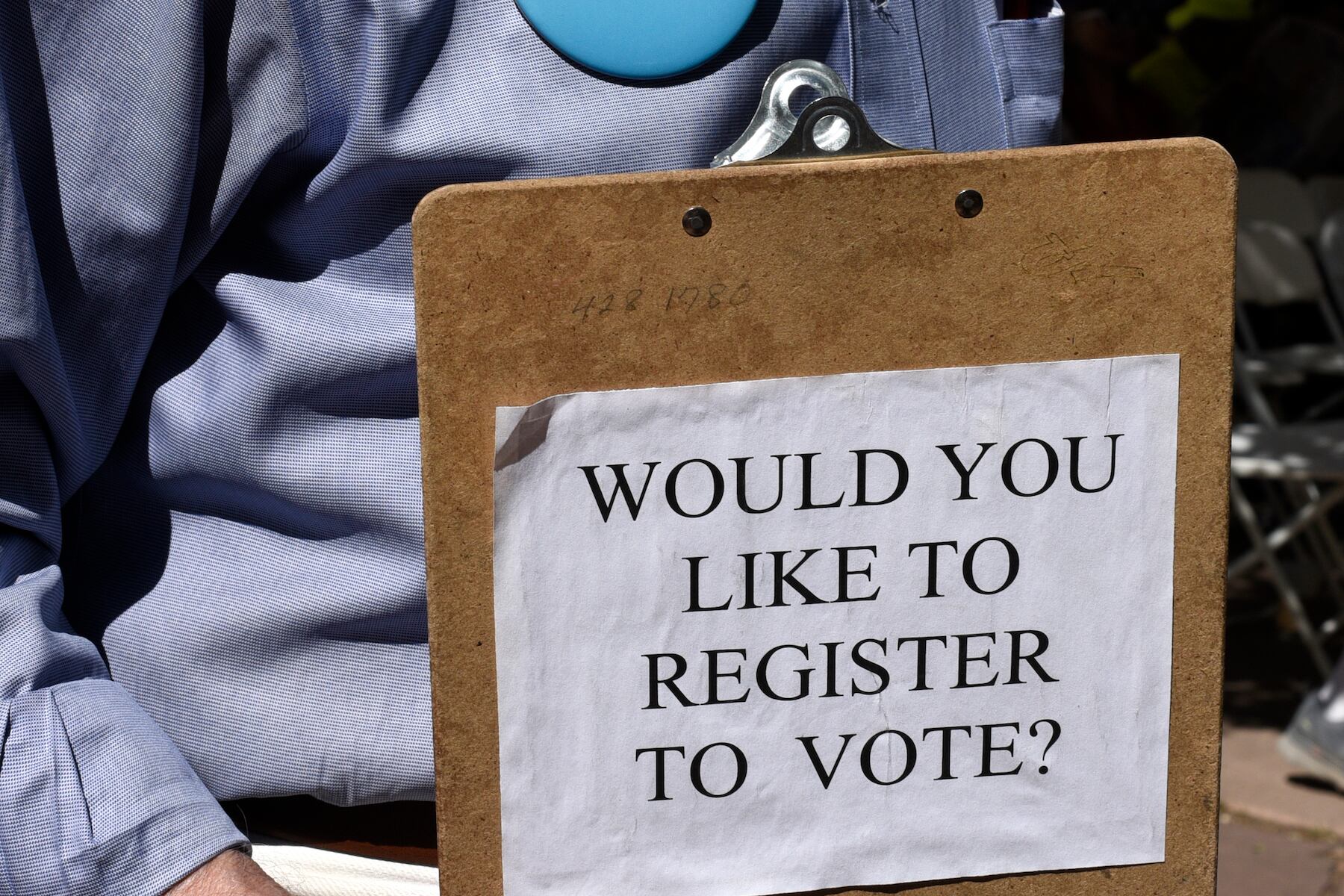 A man in a blue shirt holds a clipboard, the back of which has a page taped to it reading “Would you like to register to vote?”