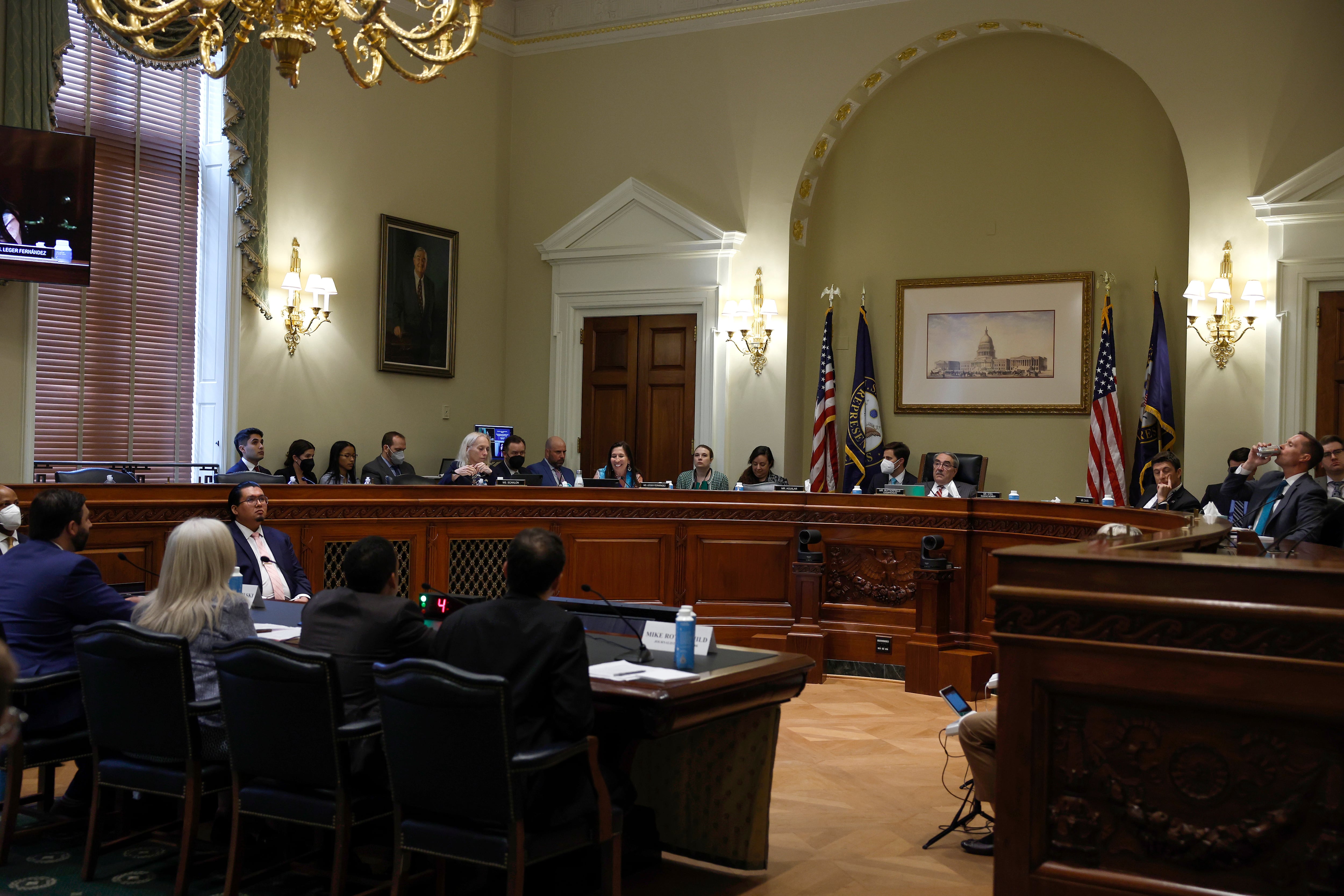 Men and women gather around a raised wooden dais, facing other men and women seated at tables in the foreground with their backs to the camera.