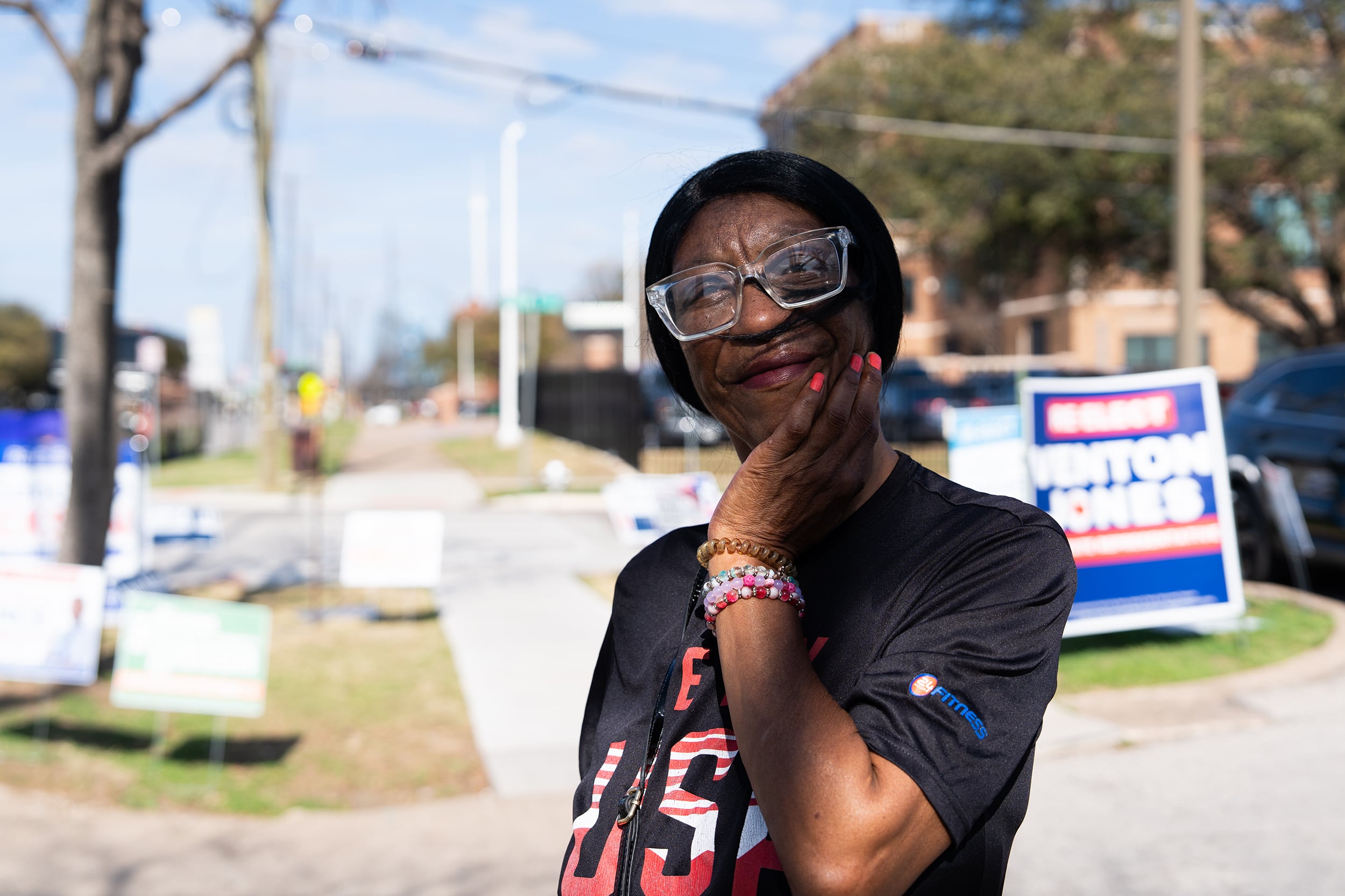 A photograph of a Black woman in a black t-shirt with "USA" in colorful letters poses for a portrait outside on a sunny day.