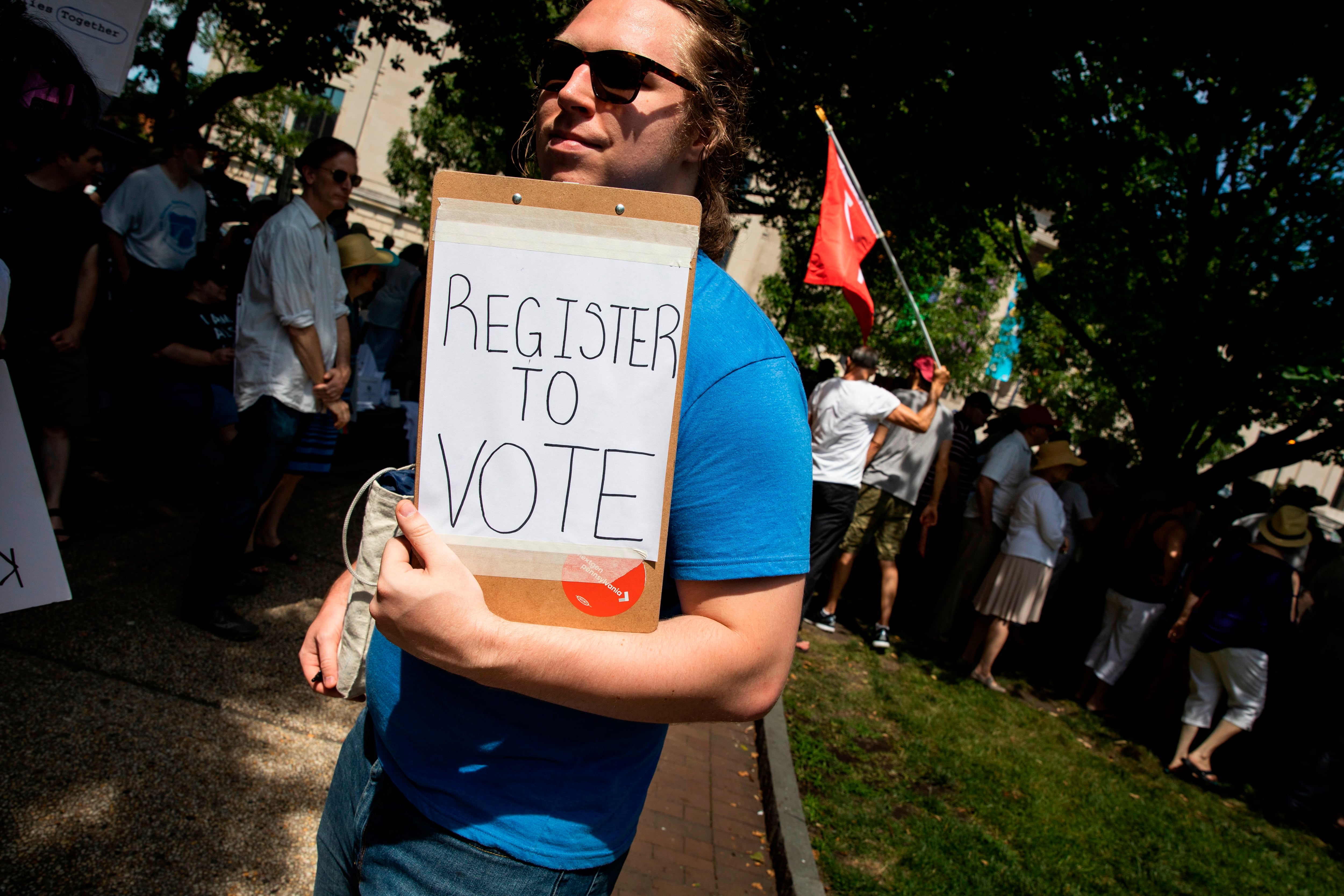 A person holds a clipboard that reads “REGISTER TO VOTE” on white paper.