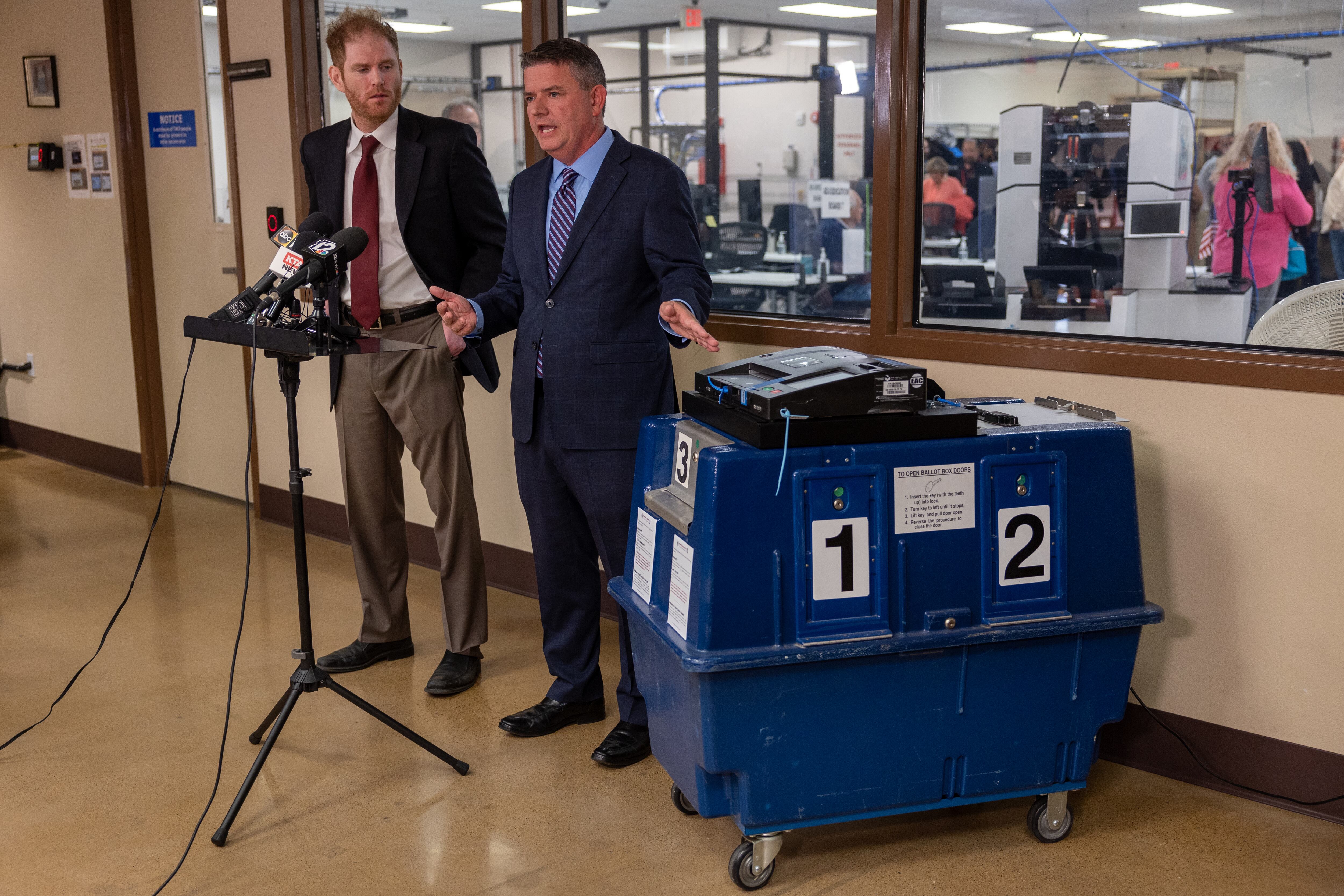 Two men in dark suits stand with their back against a wall with a viewing window set into it. One man has his hand on a voting machine.