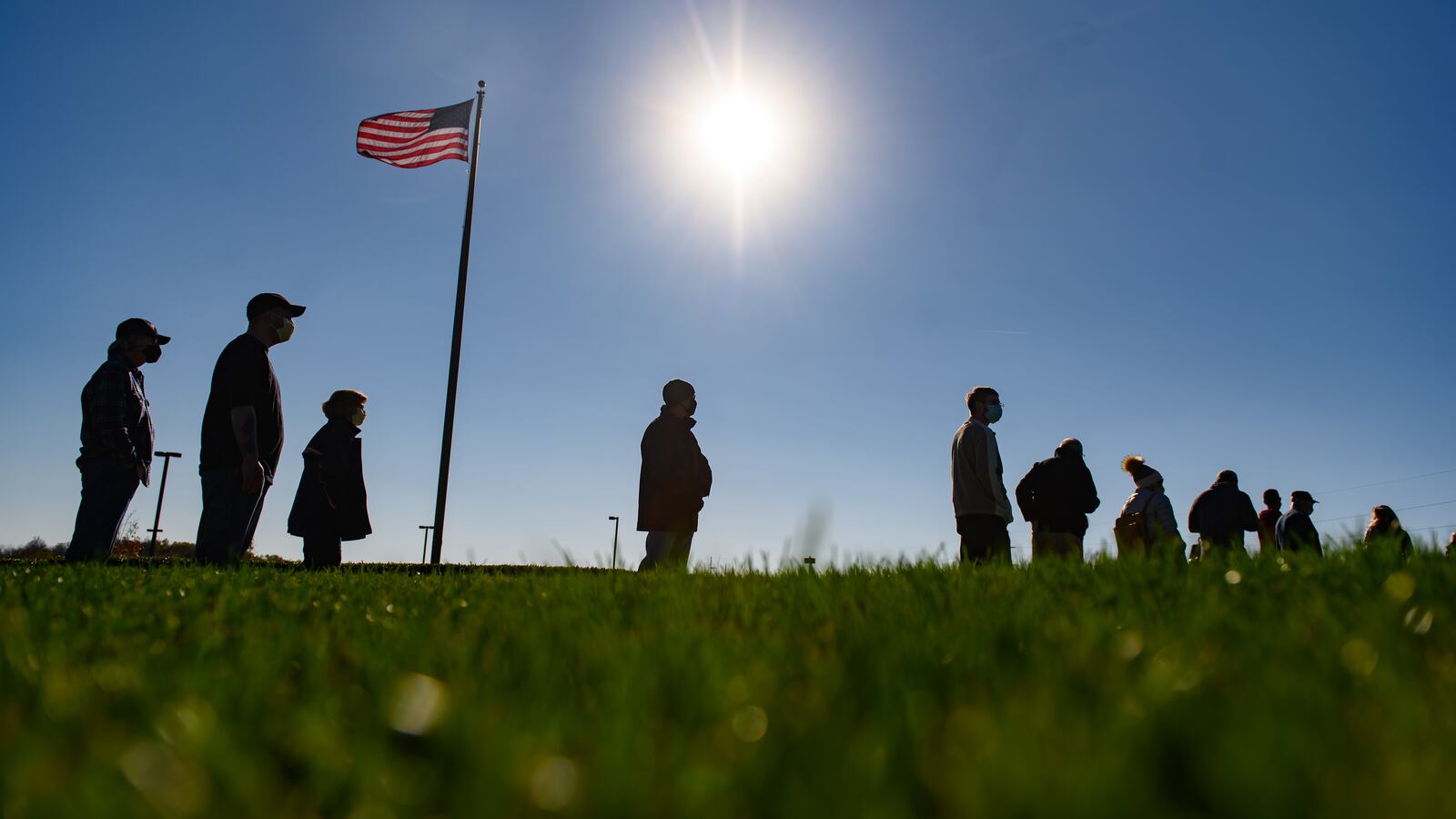 A row of people in silhouette standing on a grassy hill with a U.S. flag waving in the background