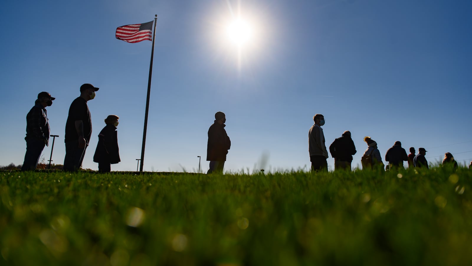 A row of people in silhouette standing on a grassy hill with a U.S. flag waving in the background