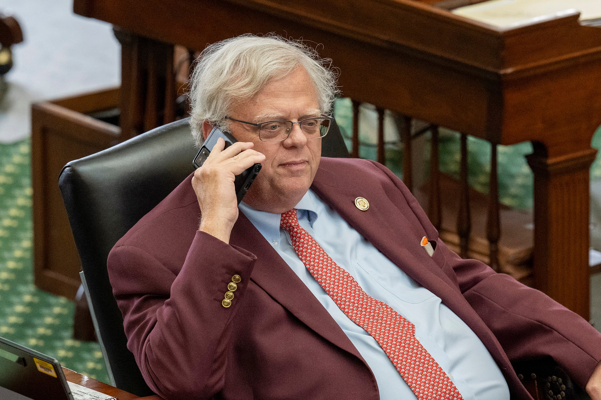 A white man in a suit sits in a black leather chair holding a cellphone to his ear in a large senate floor room.
