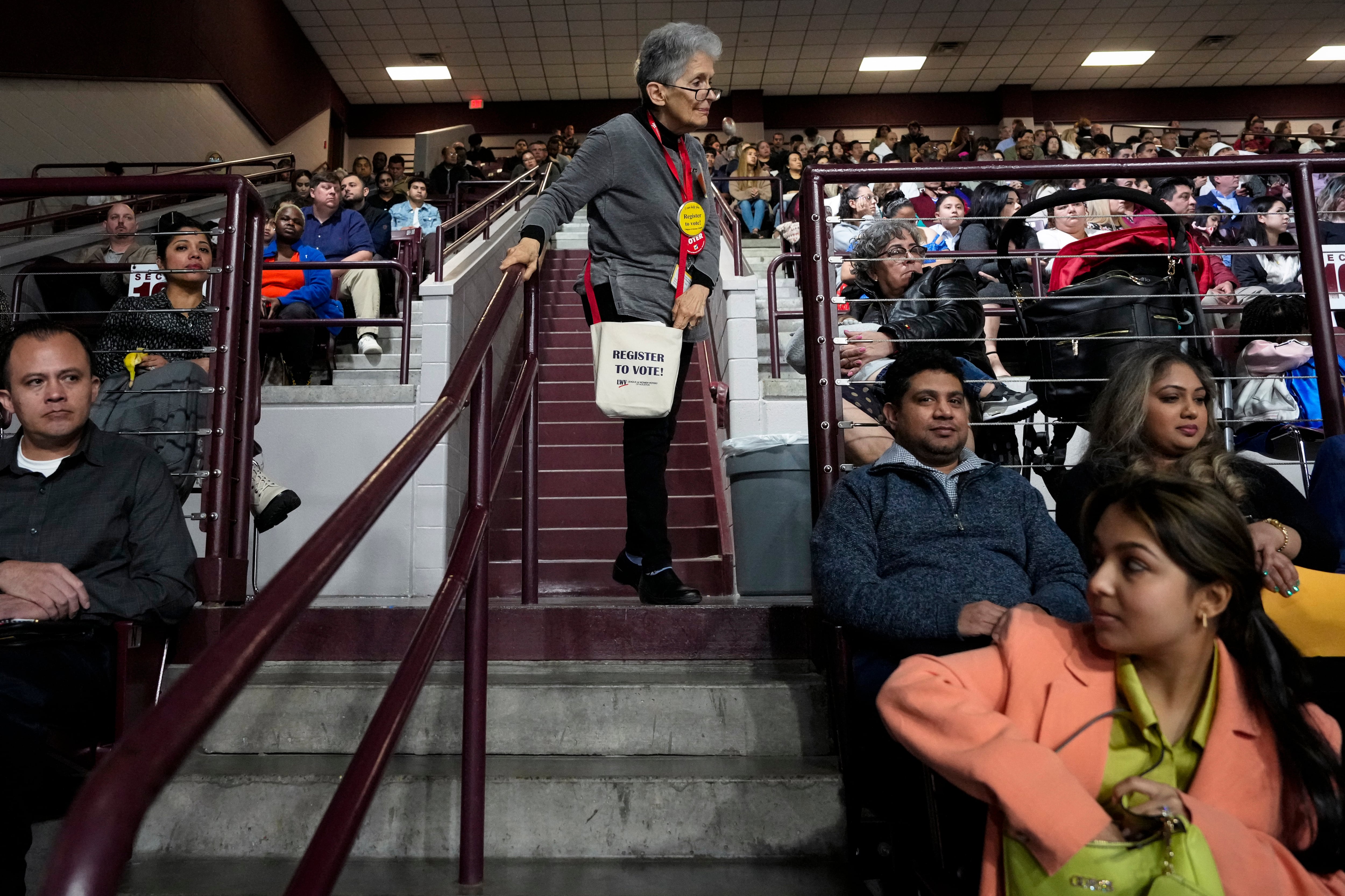 A woman in a gray top stands while many other people are seated around her.