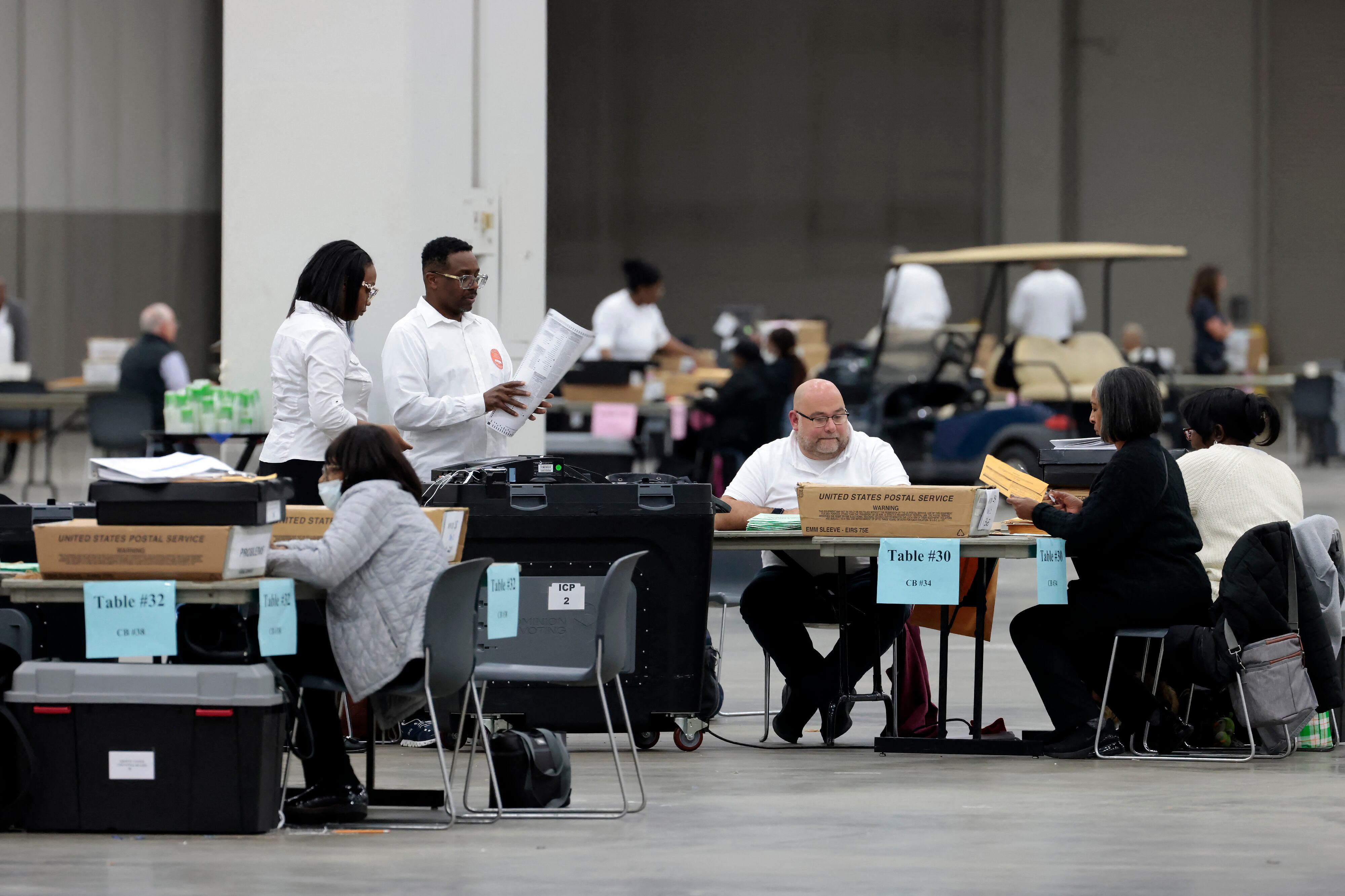 A group of adults, some standing and some sitting in a large room handling ballots.