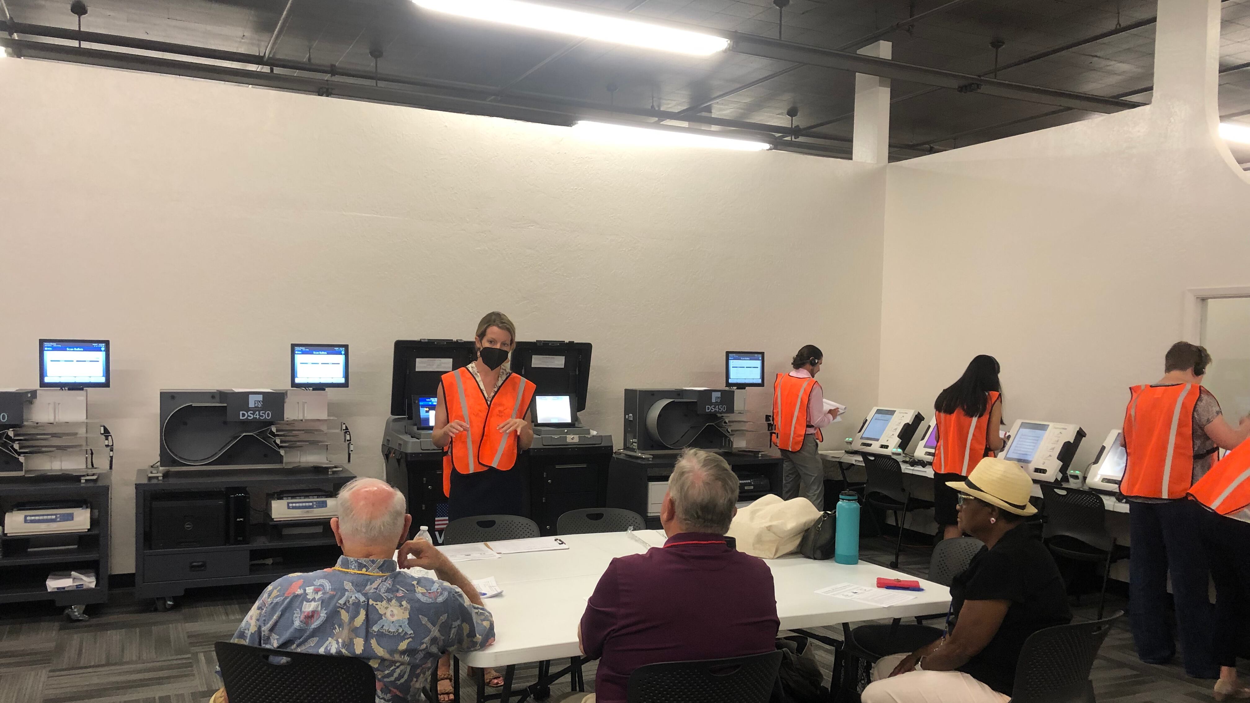 A woman in an orange vest gestures before computerized machines while three seated observers watch.