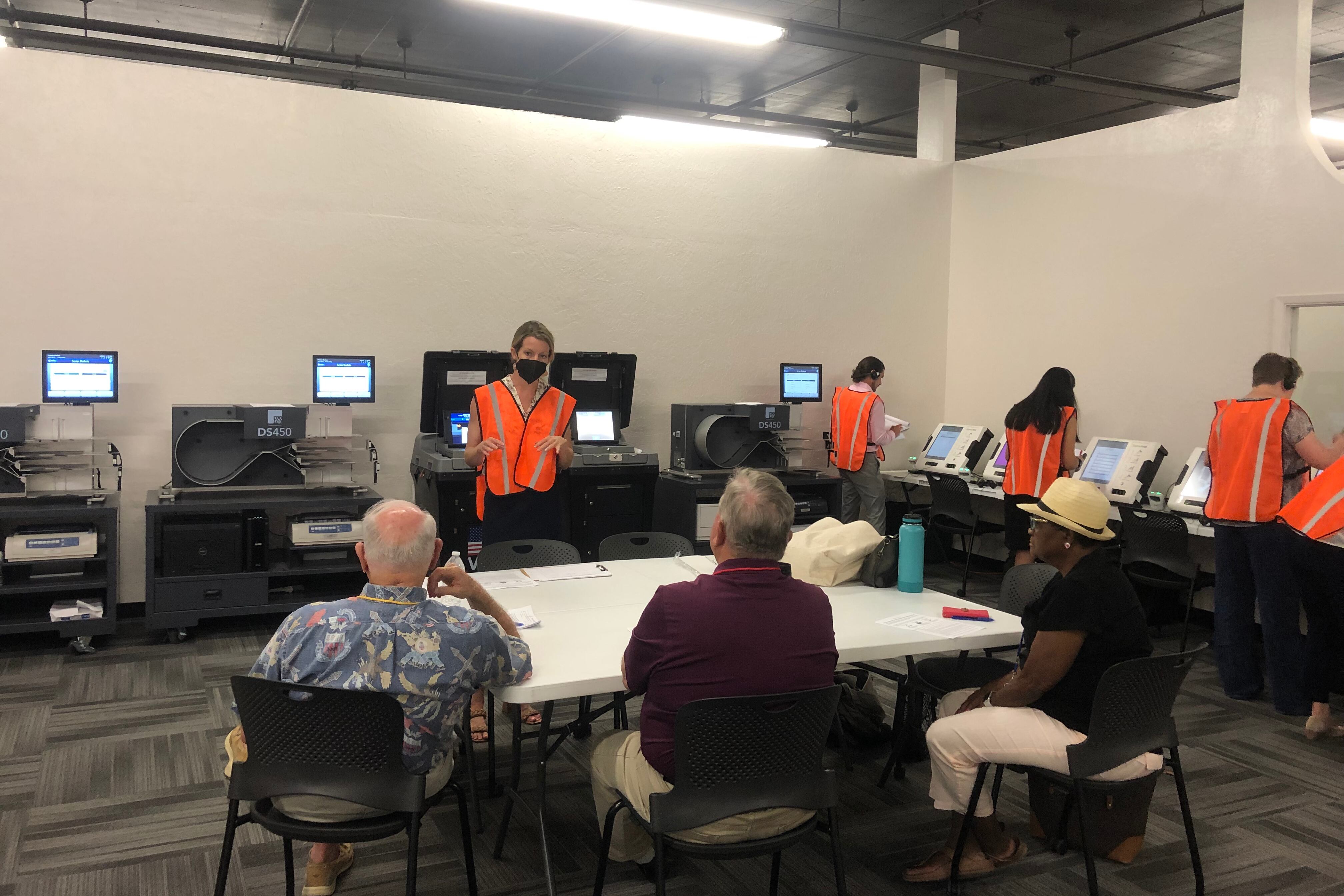 A woman in an orange vest gestures before computerized machines while three seated observers watch.