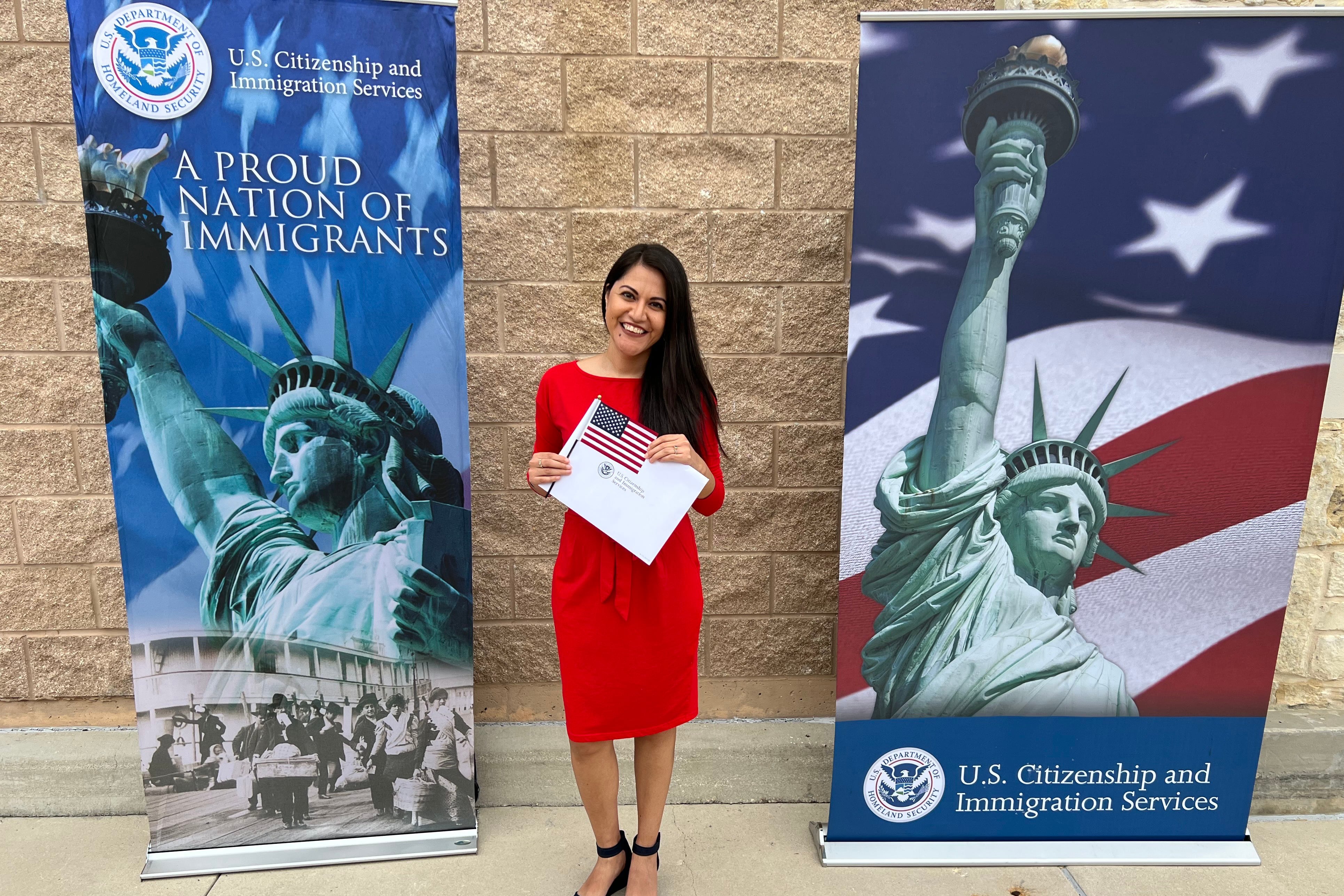 A woman with long dark hair and wearing a bright red dress holds a piece of paper and a small American flag between two banners outside of a tan stone building.