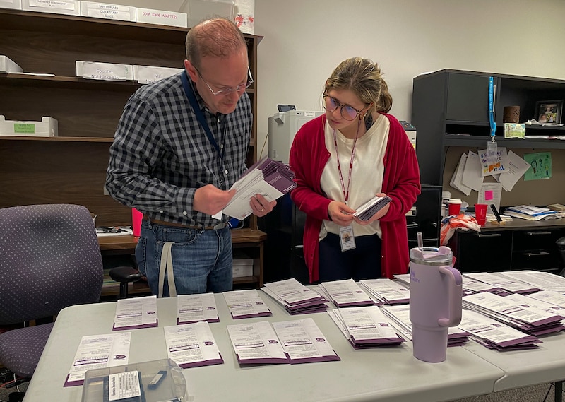 Two people stands over a table with stacks of paper across it.