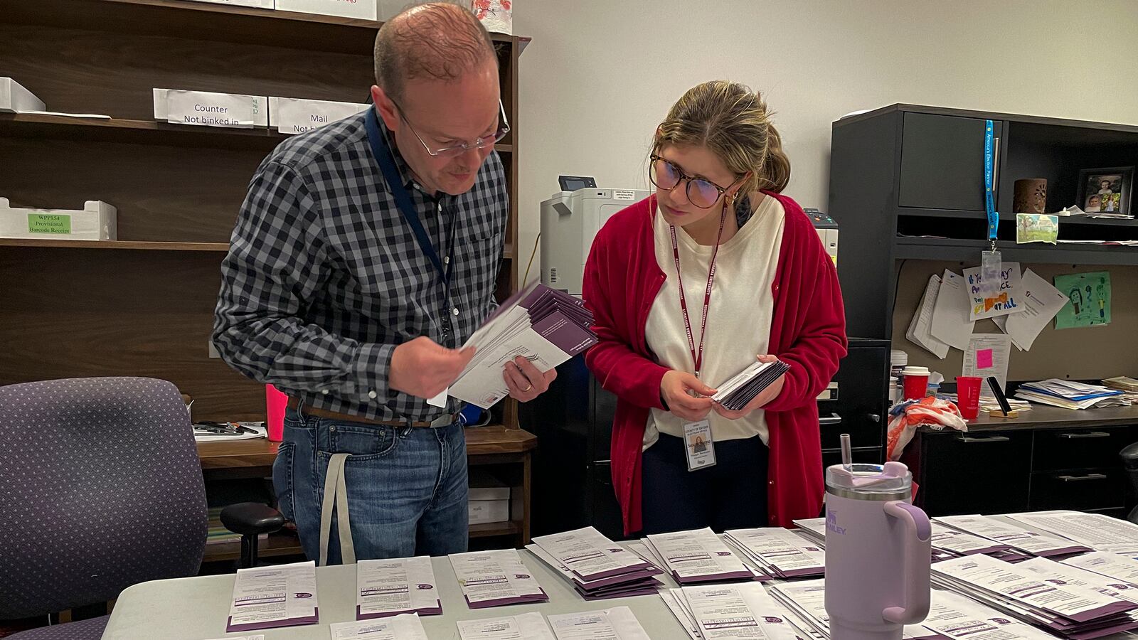 Two people stands over a table with stacks of paper across it.
