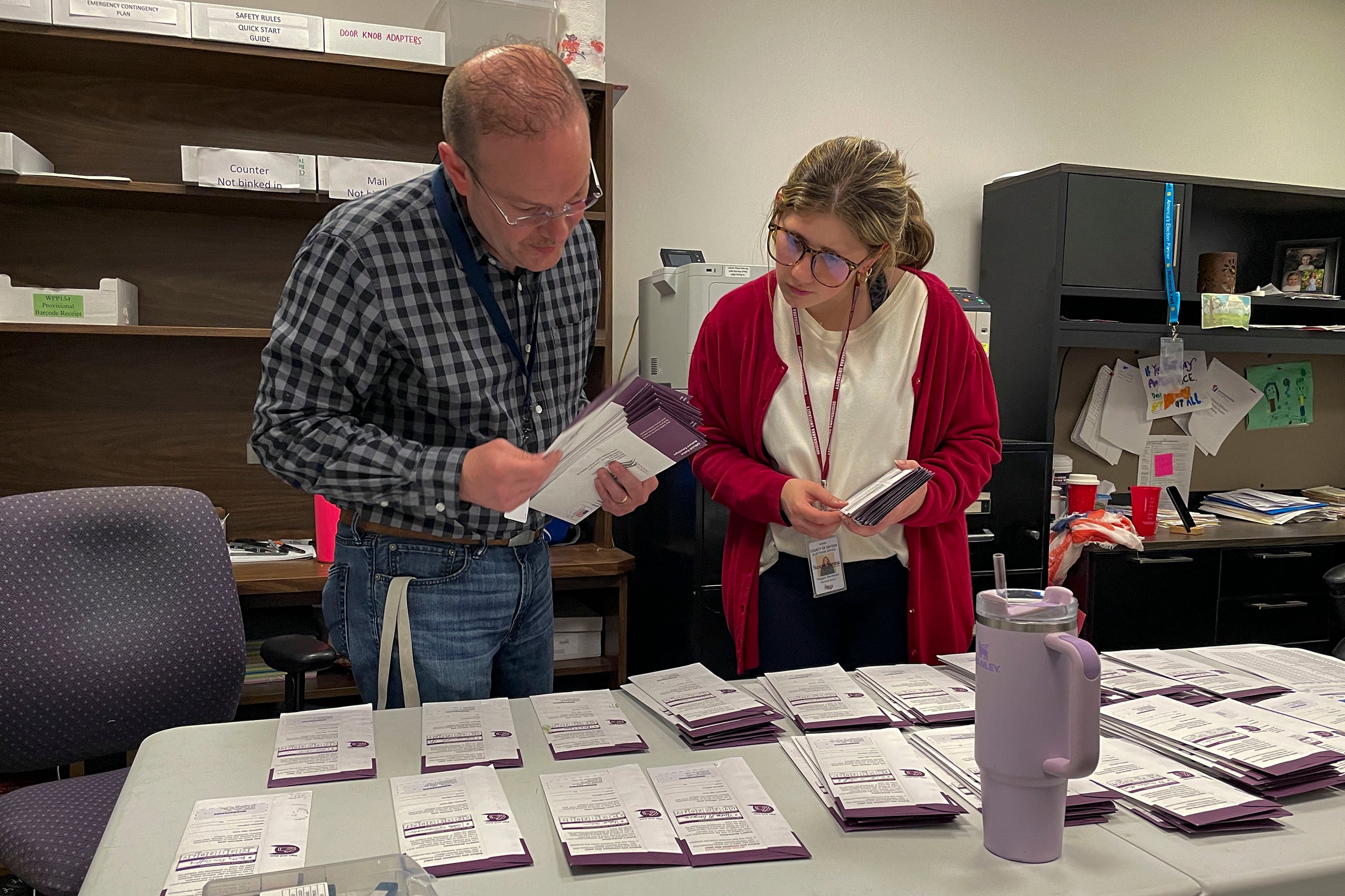 Two people stands over a table with stacks of paper across it.