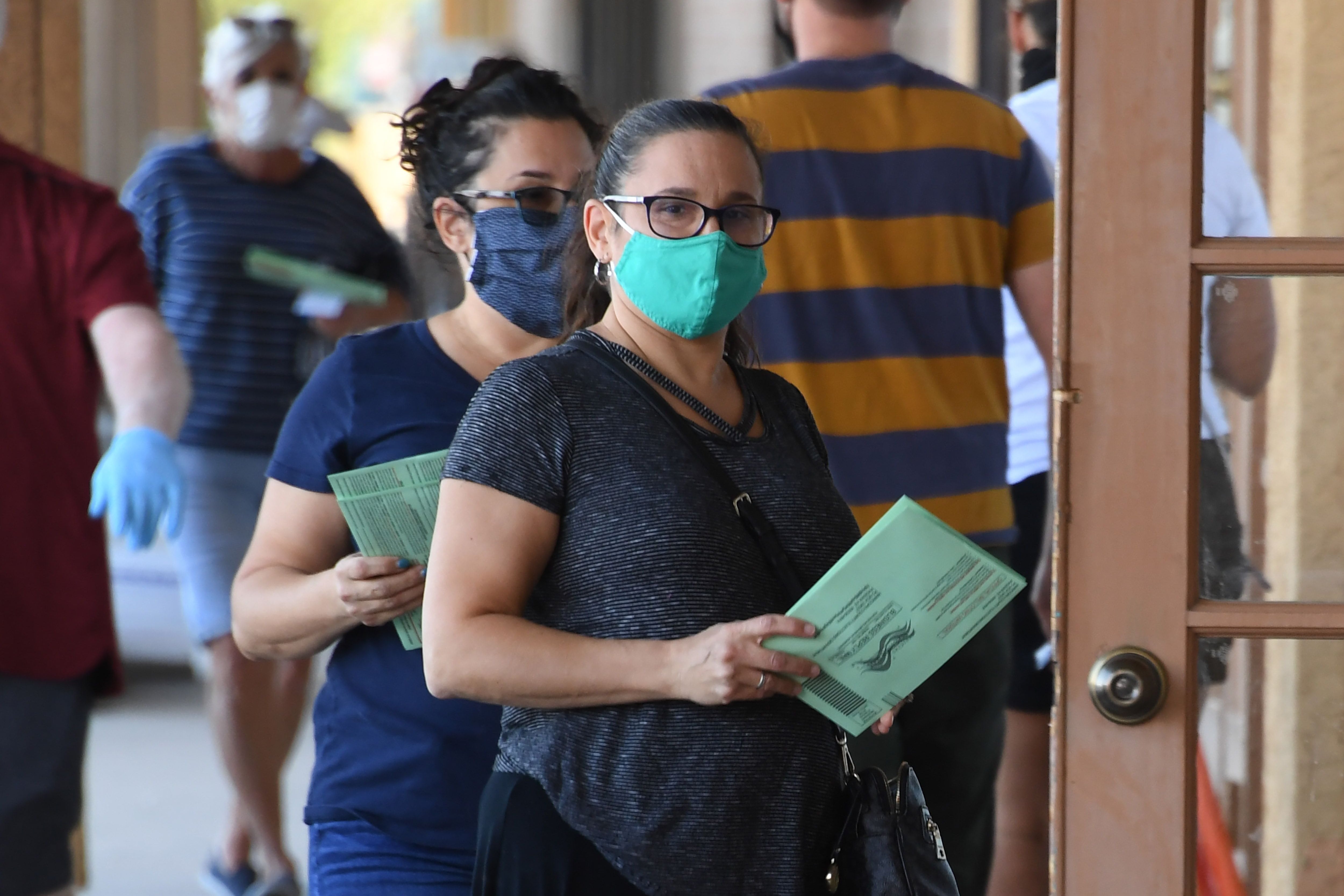 Two people with face masks wait outside door of polling place with green ballot envelopes in their hands.