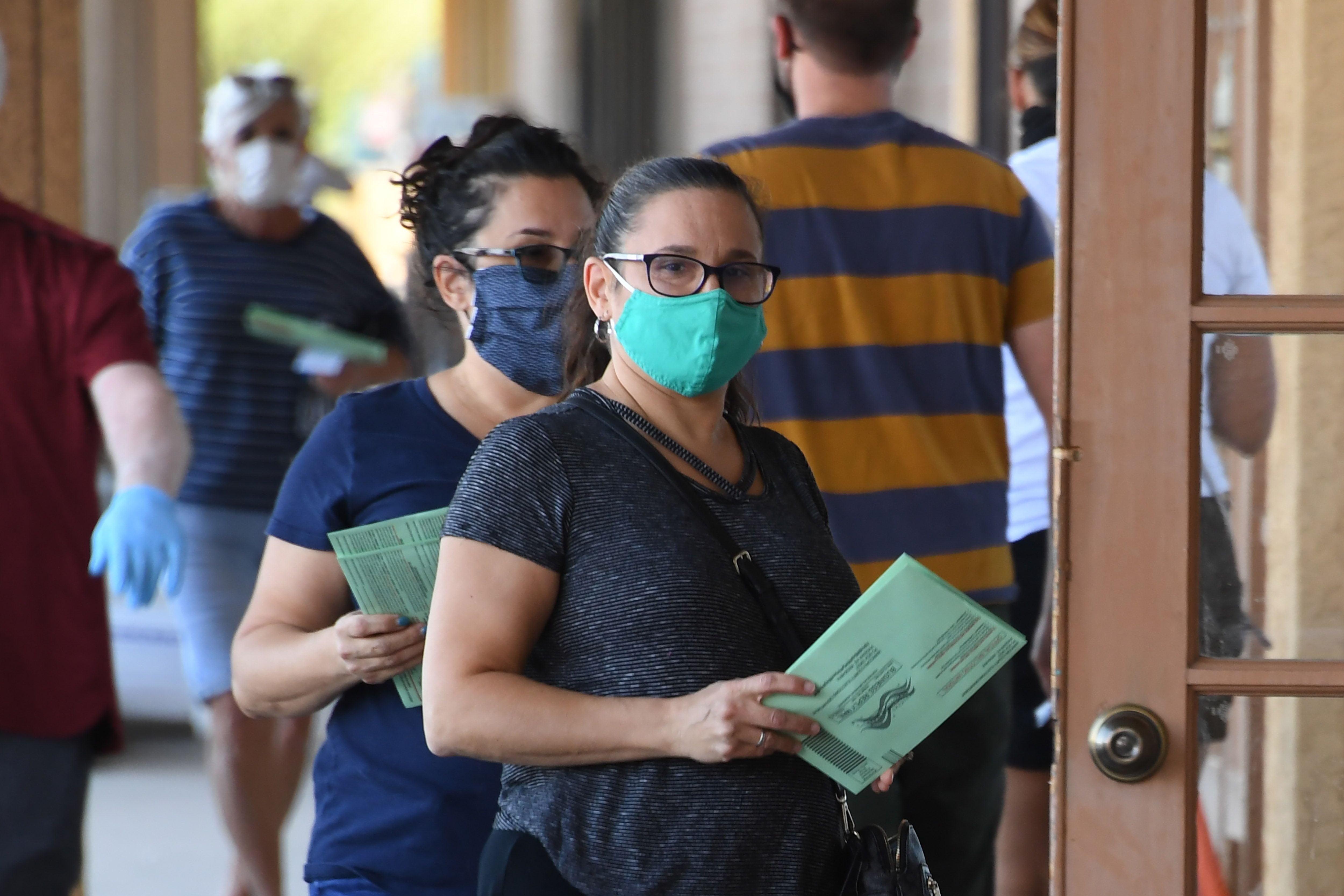 Two people with face masks wait outside door of polling place with green ballot envelopes in their hands.