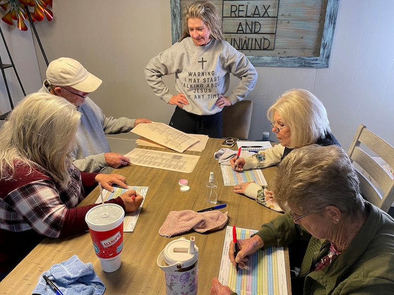 A photograph of a white woman in a sweater standing next to a group of four white people sitting at a table working on a piece of paper.