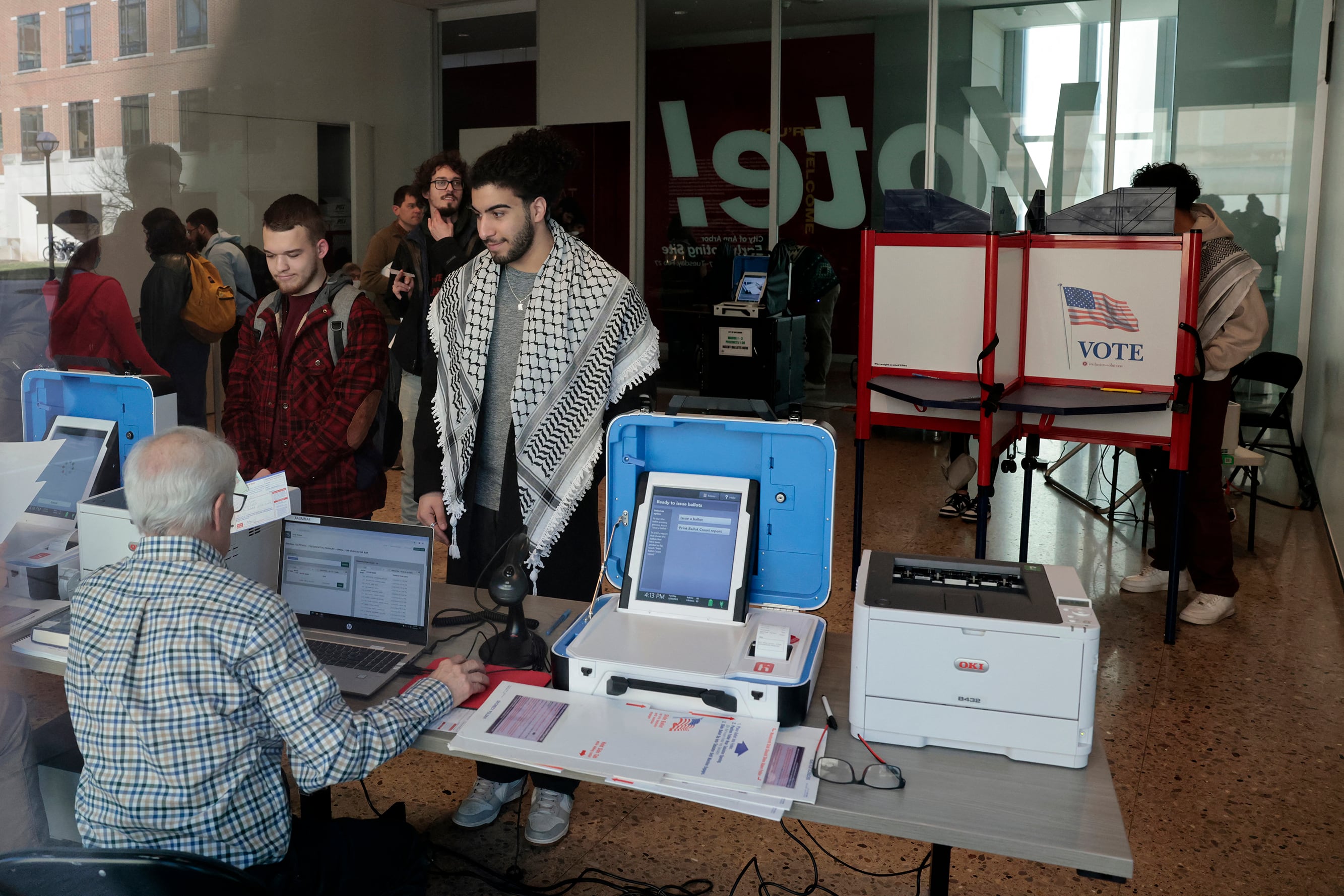 Two people stand in front of a desk with voting equipment and a person sitting at the desk. one person is voting in the background.