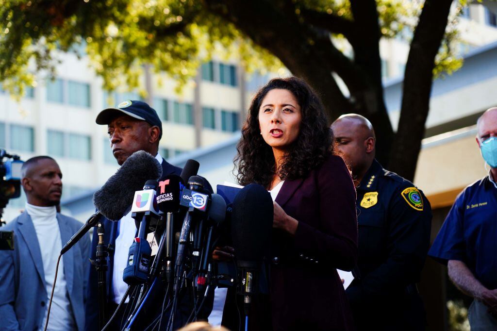 A woman with dark curly hair stands before a bank of microphones, with a police officer and other men in the background. 