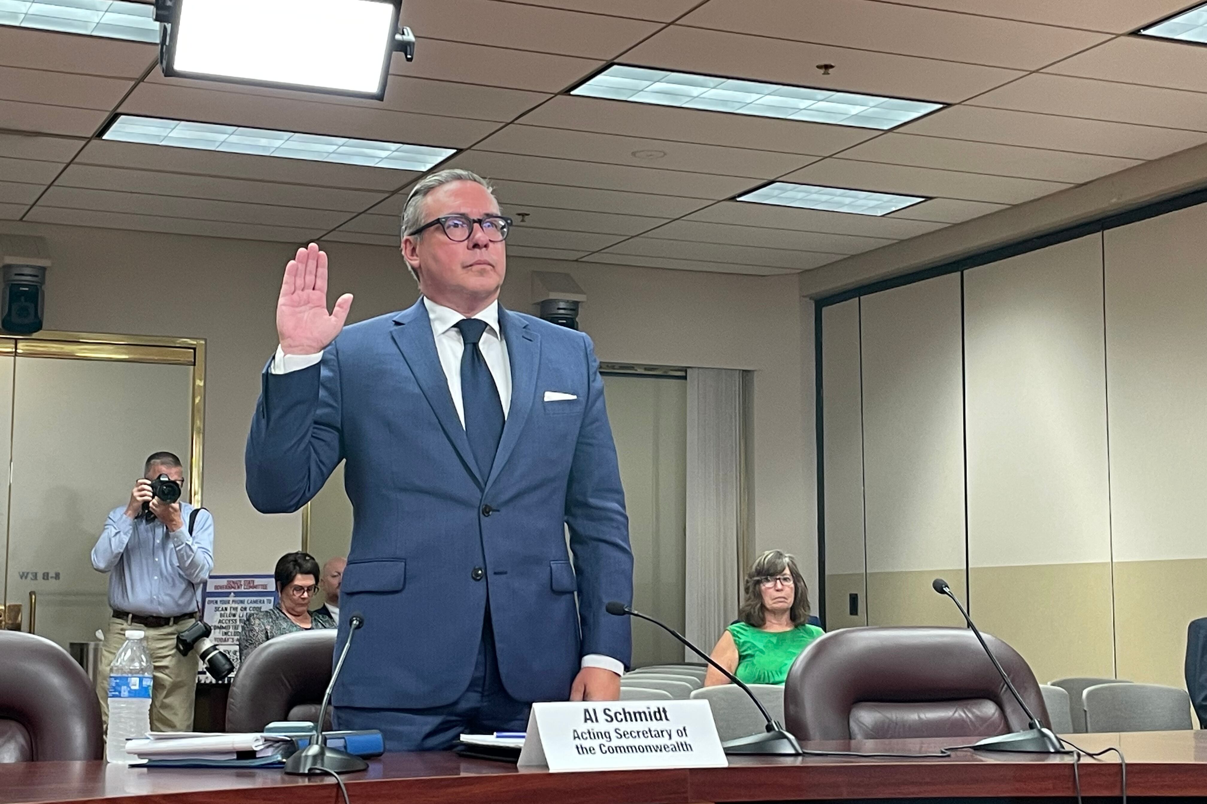 A man stands at a table in a hearing room with his right hand raised.