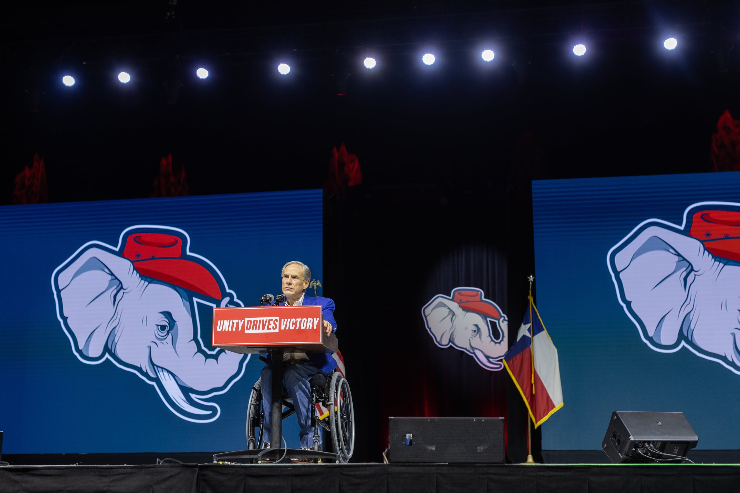 A photograph of a white man wearing a suit sitting in a wheelchair on a stage speaking into a microphone.